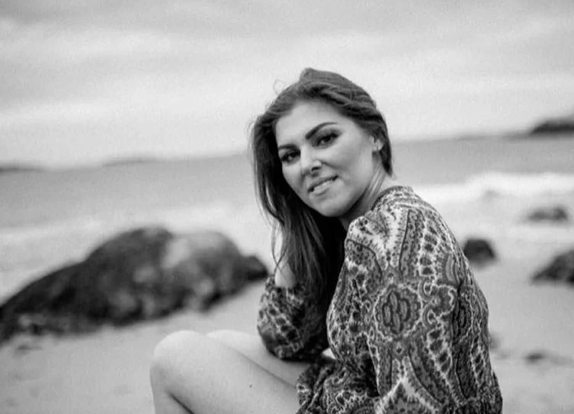 Young woman sitting on the beach, smiling, with rocks and water in the background.