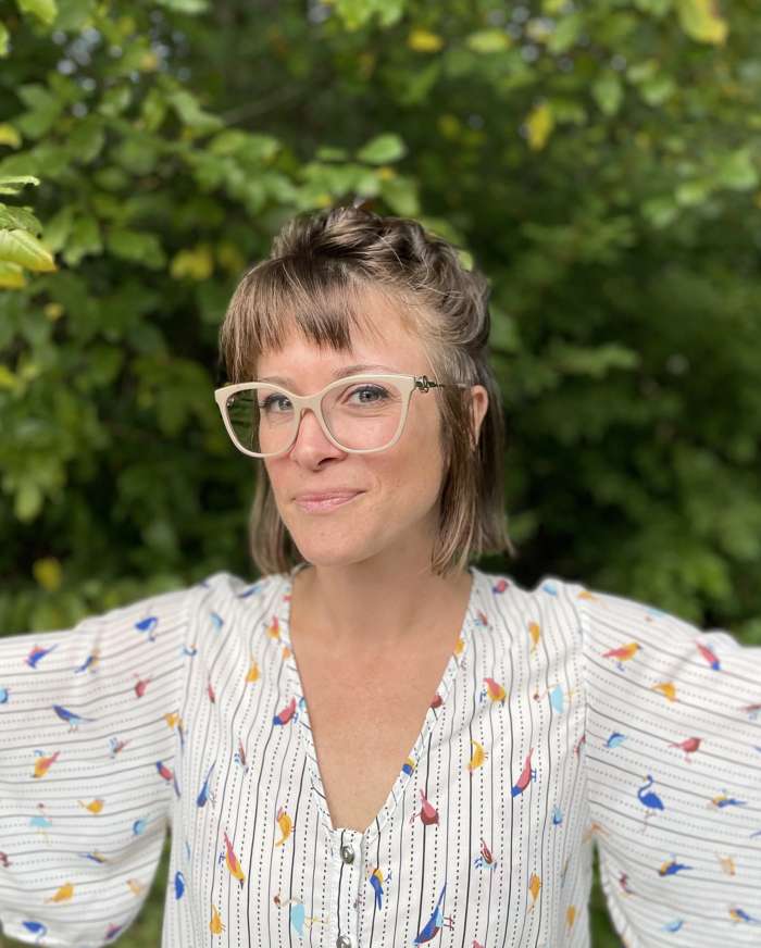 A woman with short brown hair, wearing glasses and a colorful bird-patterned blouse, standing outdoors in front of green foliage.