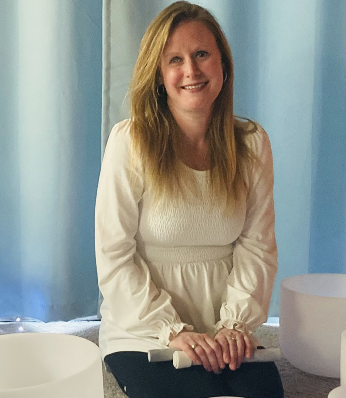 A woman with long blonde hair, smiling, sitting on the floor with her hands resting on a white object, wearing a white long-sleeved top, with blue curtains in the background and crystal bowls around her.