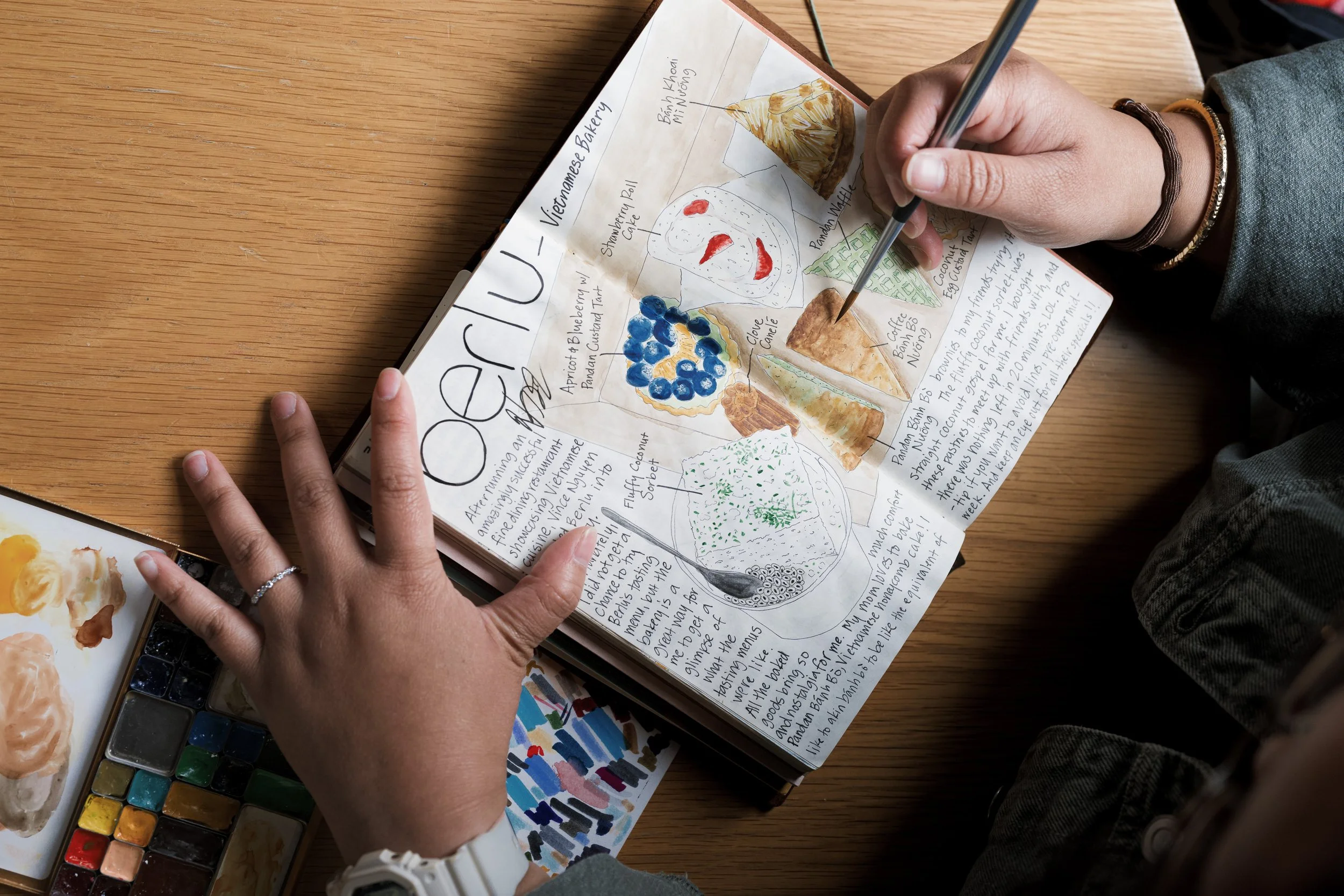 A person is creating a colorful travel journal page with watercolor illustrations of Vietnamese desserts. The page features drawings of coconut pudding, blueberry pastry, and pandan cake, with handwritten notes describing each item on a wooden table. Art supplies, including a watercolor palette and a paintbrush, are also visible.