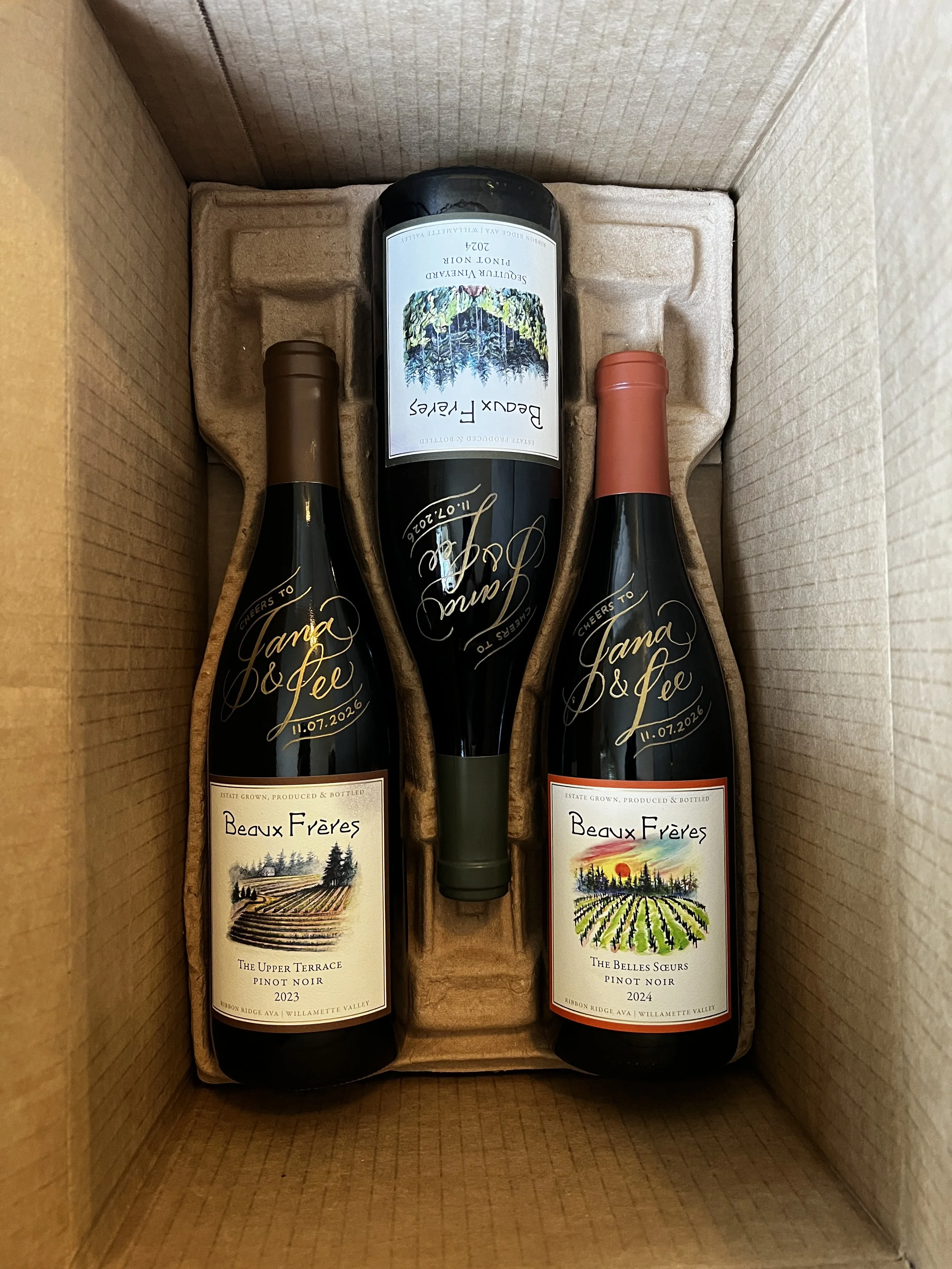 Inside a cardboard box, two bottles of Beaux Frères Pinot Noir and one bottle of Sourture Vineyard Pinot Noir. The Beaux Frères bottles' labels show landscape artwork, with one from 2023 and the other from 2024. The middle bottle's label features a forest scene with colorful trees and a sunset.