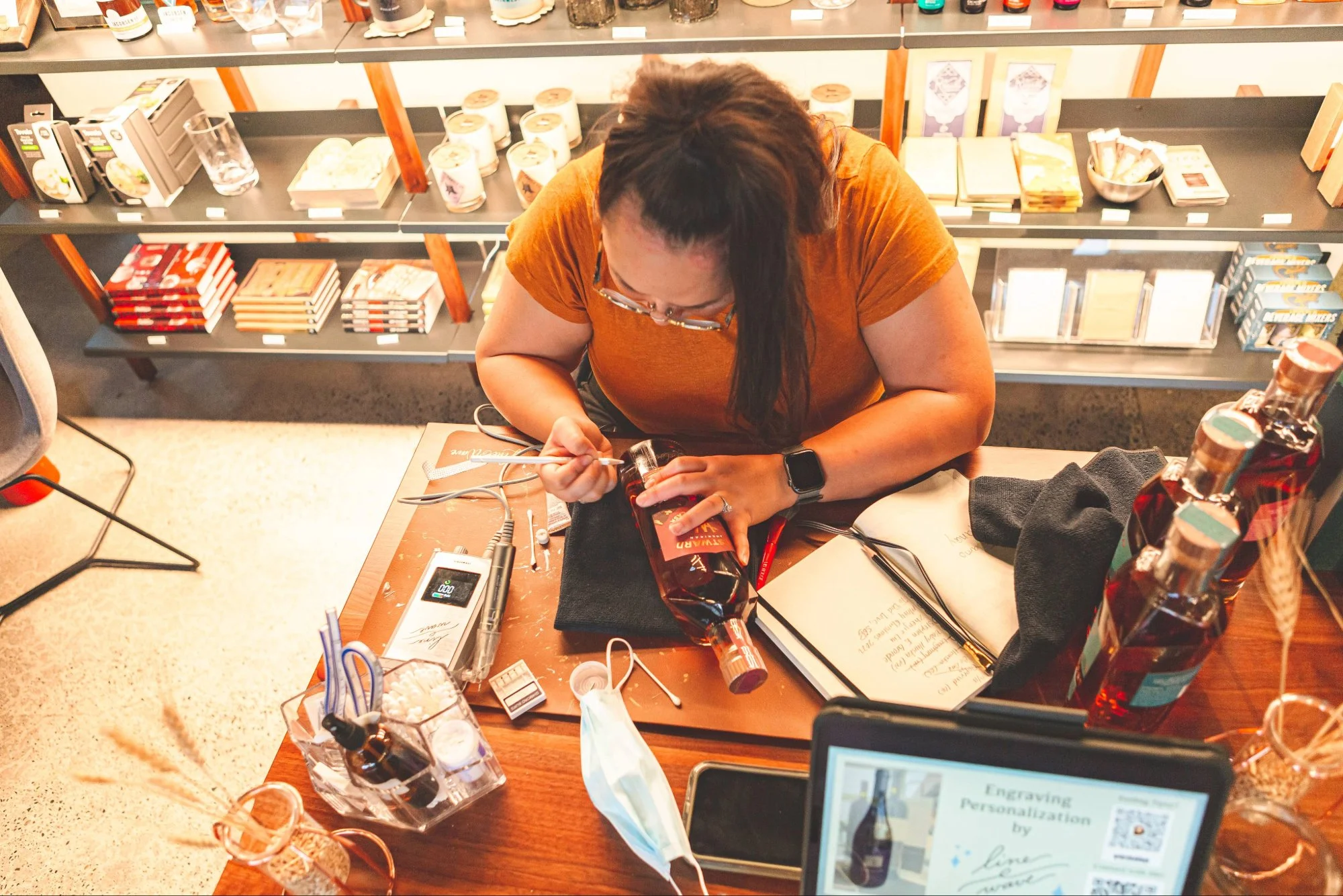 A woman with long dark hair and glasses, wearing an orange shirt, is sitting at a desk in a bookstore or library. She is writing on a package with a pen. The desk has various items, including a digital device, papers, and a face mask. Behind her are shelves filled with books and other products.