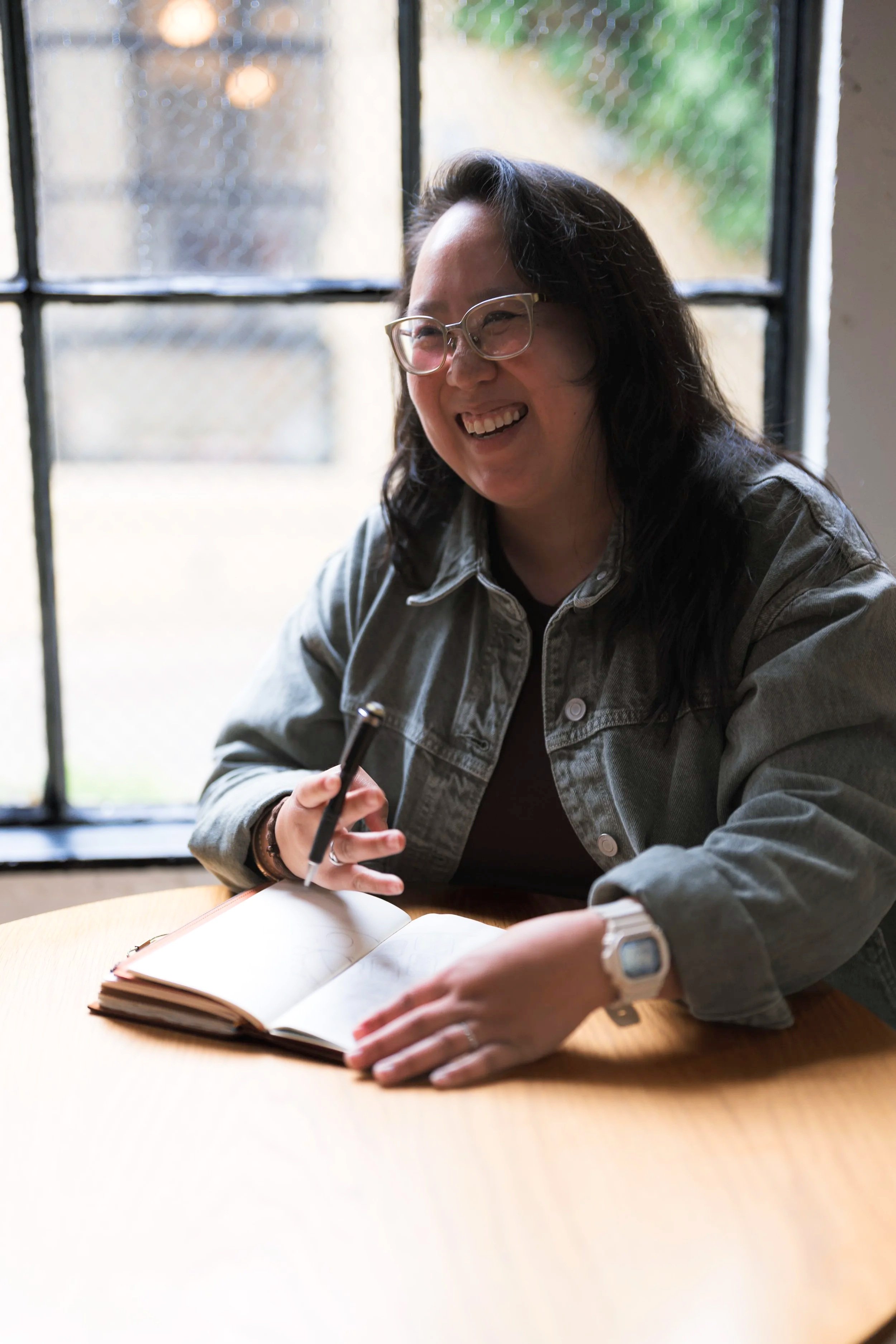 A woman with glasses and dark hair, smiling while writing in a notebook, sitting at a wooden table by a window.