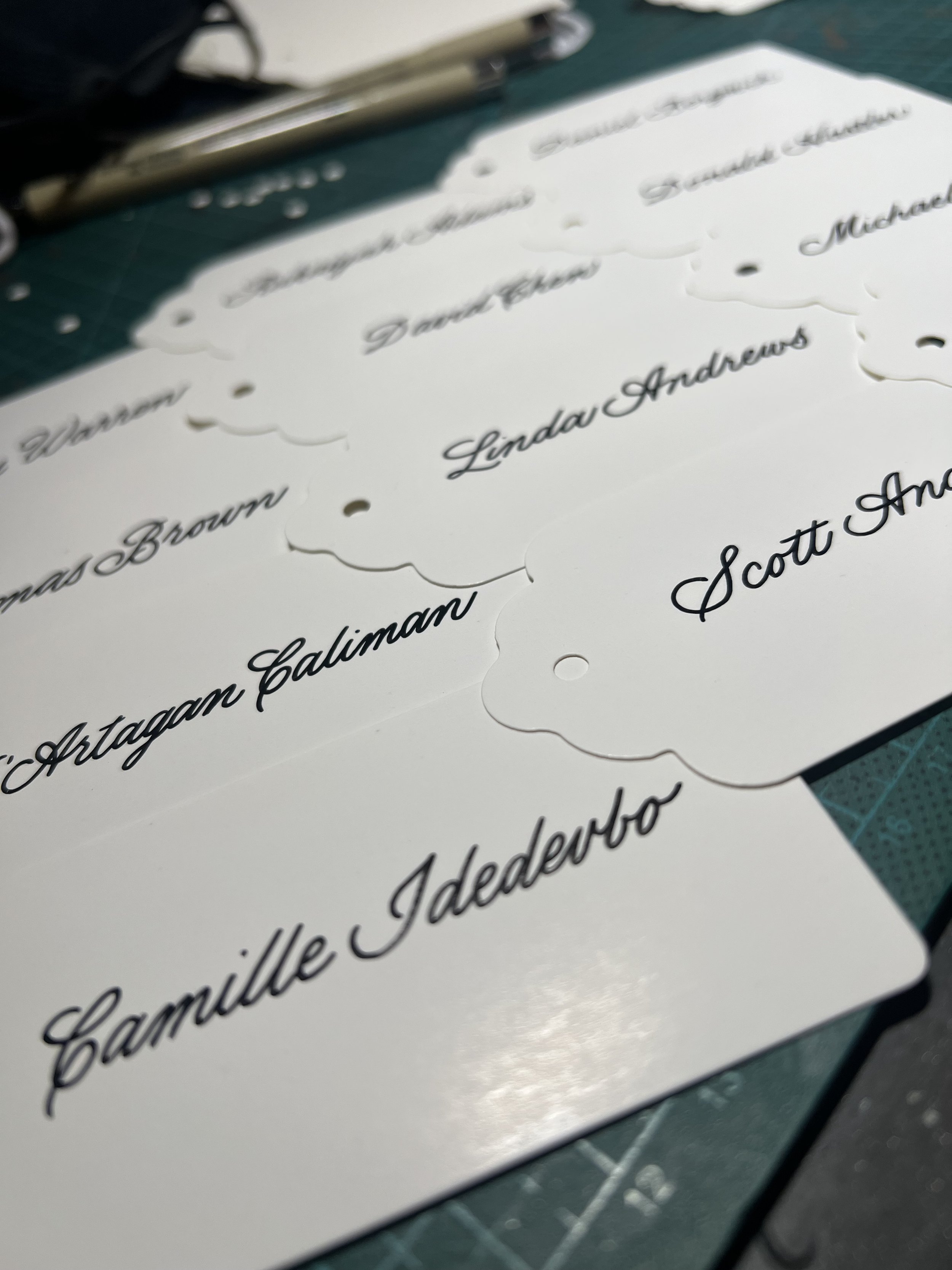 Close-up of several white paper labels with black cursive writing of different names, scattered on a green cutting mat with a pair of scissors and other materials in the background.