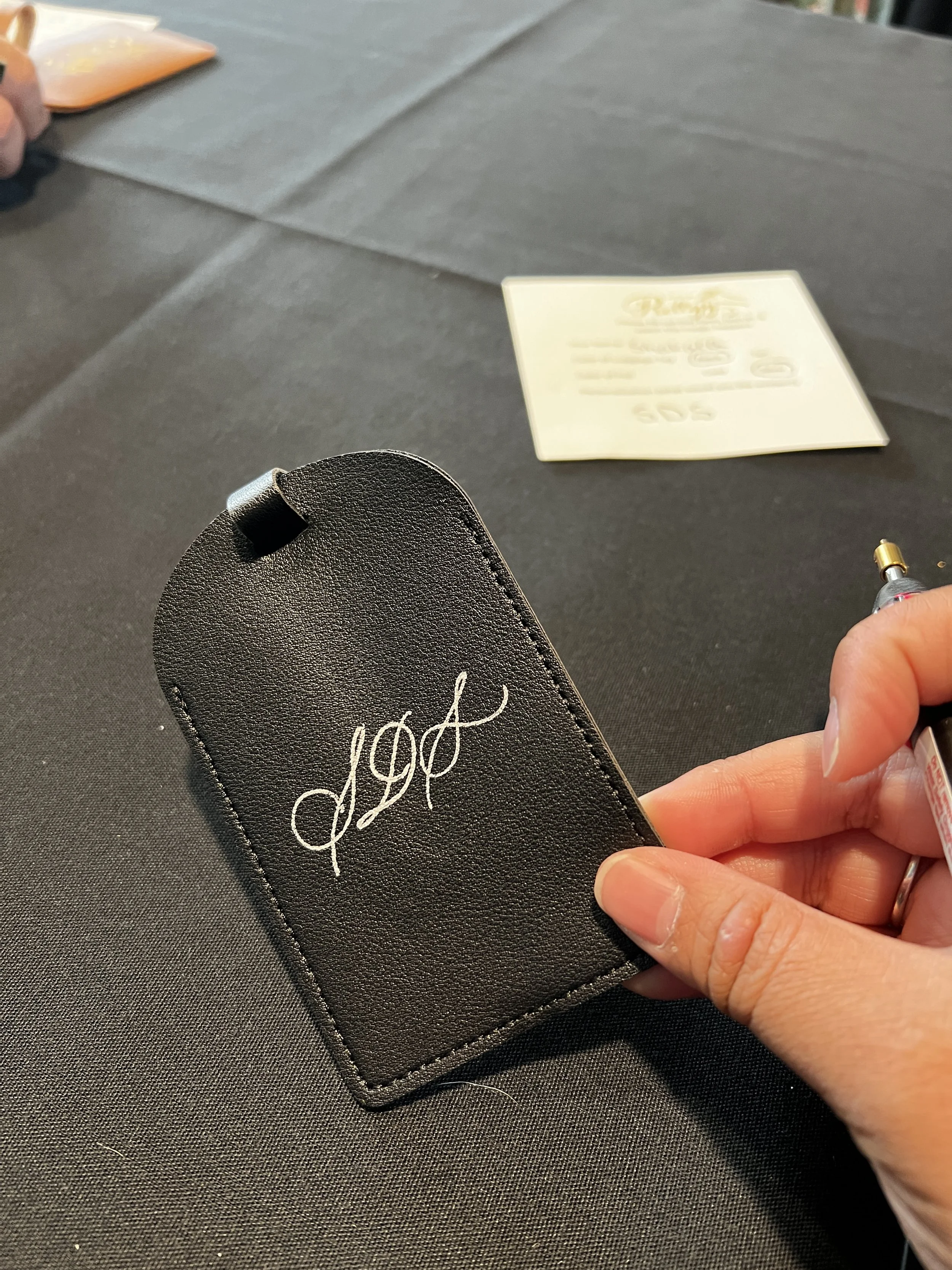 A hand is holding a pen and writing on a black tablecloth. There is a black object with white cursive text that appears to be a logo or monogram. In the background, there are some blurred papers or napkins and a printed sheet of paper on the table.