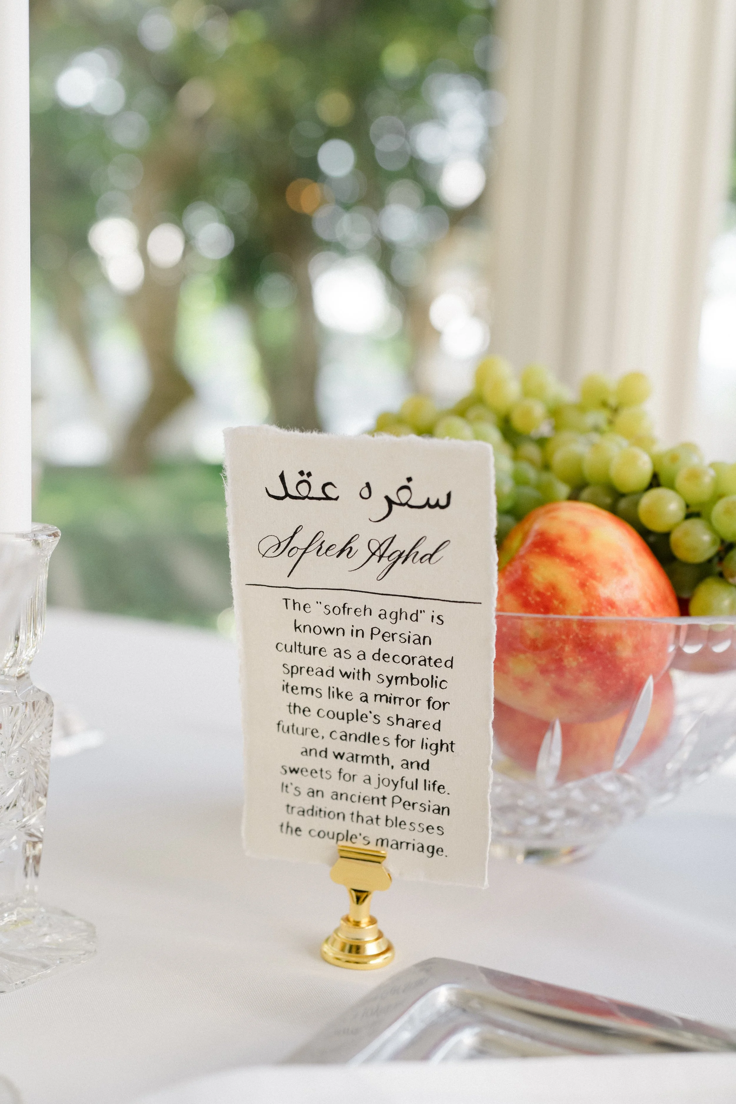 A display of a card with information about the Persian fruit, Sofreh Aghd, featuring grapes and an apple, set on a table with a white tablecloth. The background includes a window with a view of a green outdoor area.