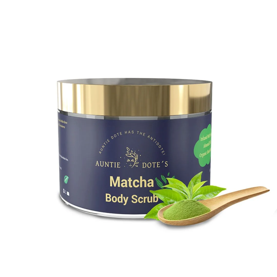 A dark blue container of Auntie Dote's Matcha Body Scrub with a gold lid, positioned beside a wooden spoon filled with green matcha powder and green leaves.