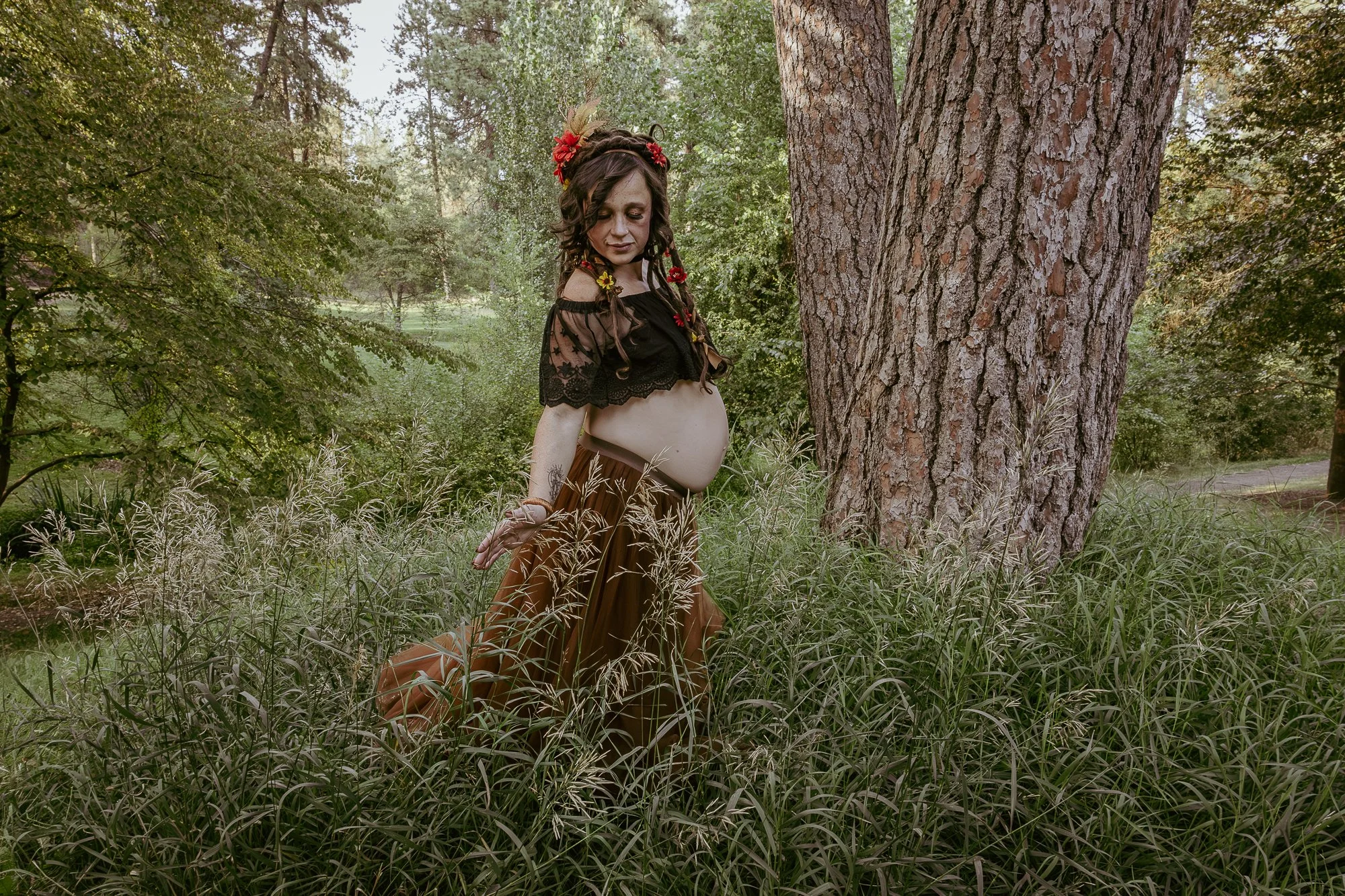 Pregnant woman in a forest setting, wearing a black lace top and long brown skirt, with flowers and leaves in her hair, standing near a large tree trunk.