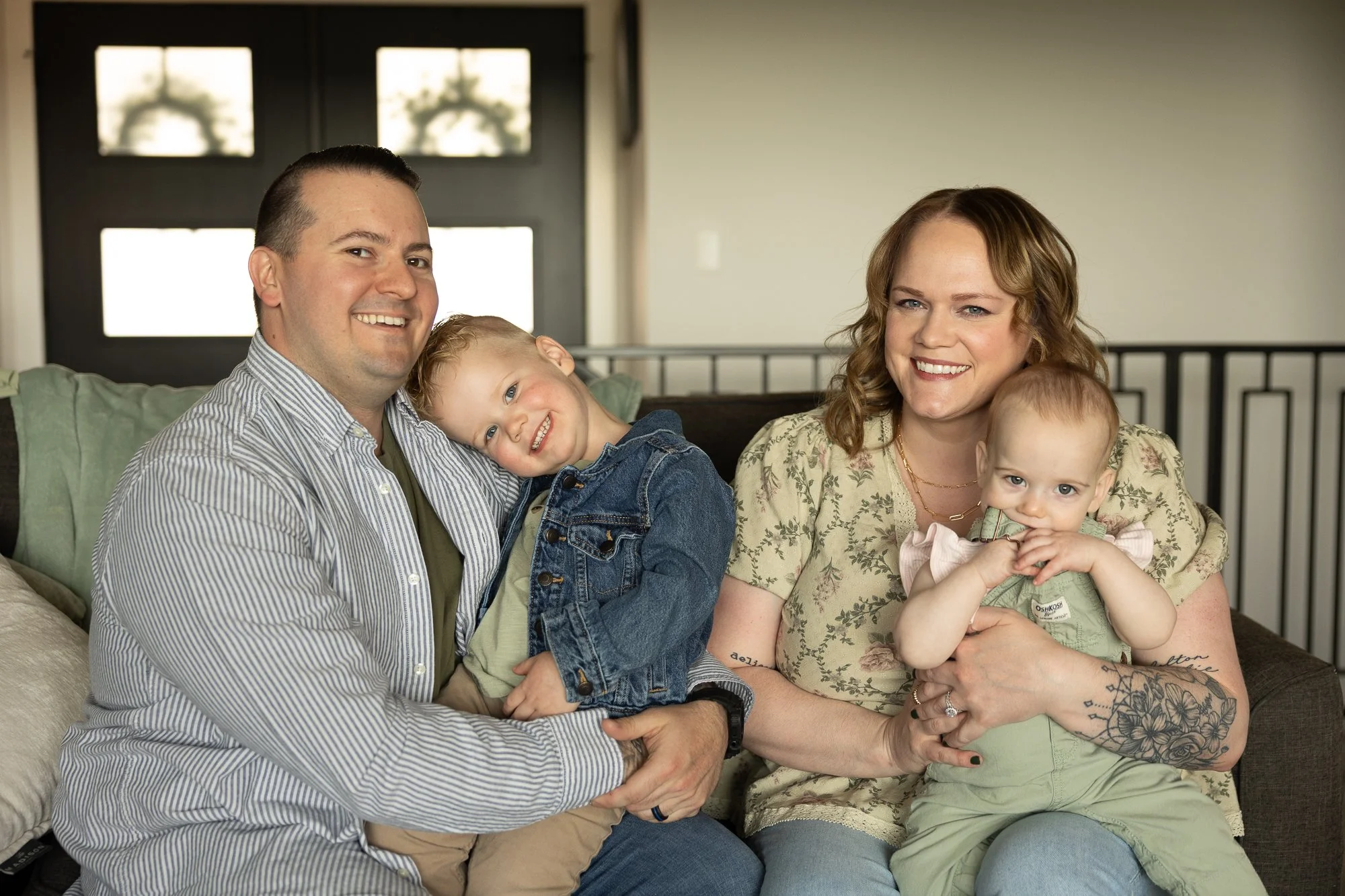 A family of four sitting on a couch inside a home, smiling at the camera. The father is on the left, wearing a striped shirt, with a young boy sitting on his lap. The mother is on the right, holding a young girl, both smiling warmly. The background features a black door with windows.
