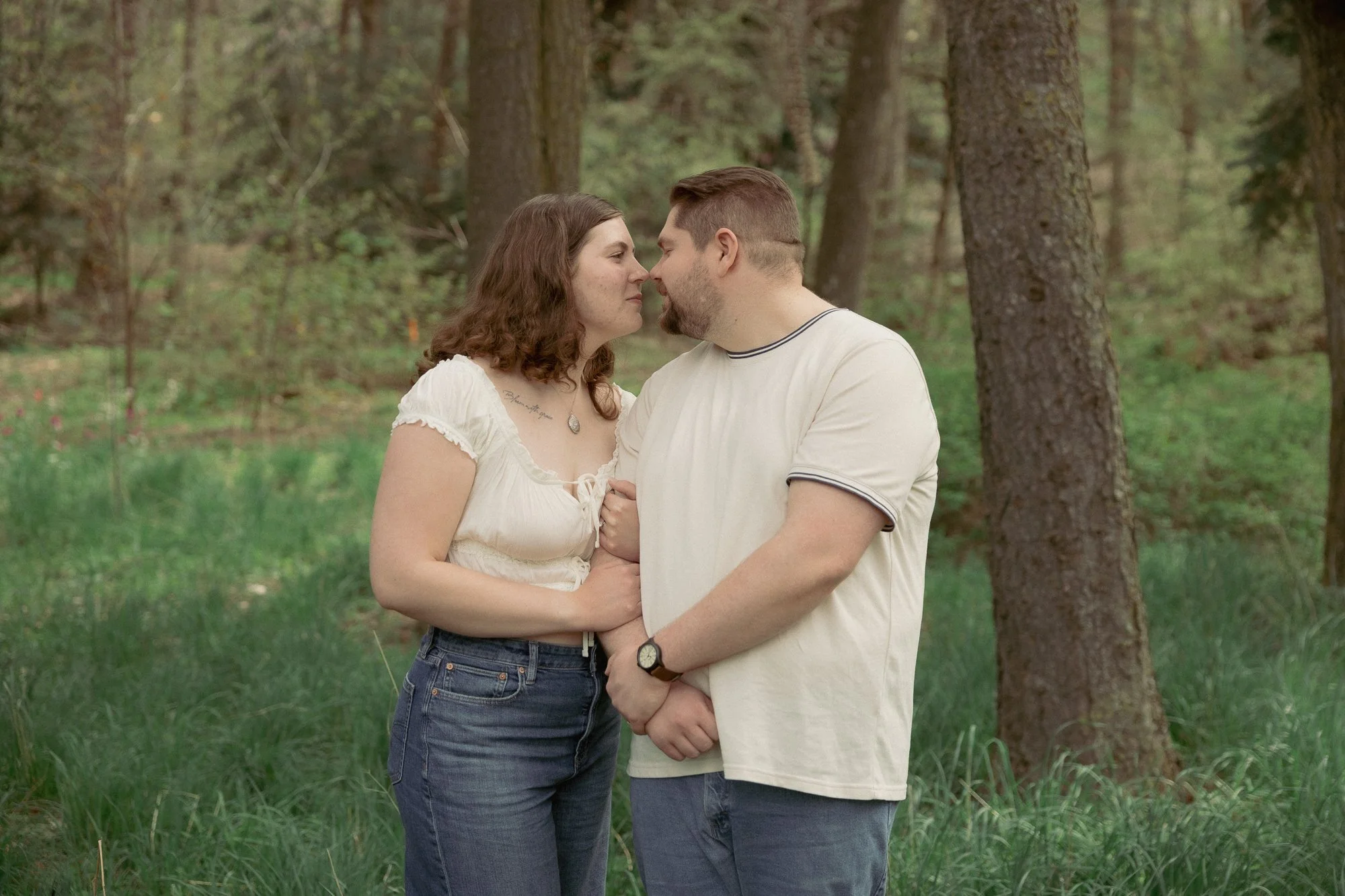 Couple standing in a grass field surrounded by trees gently touching their noses together