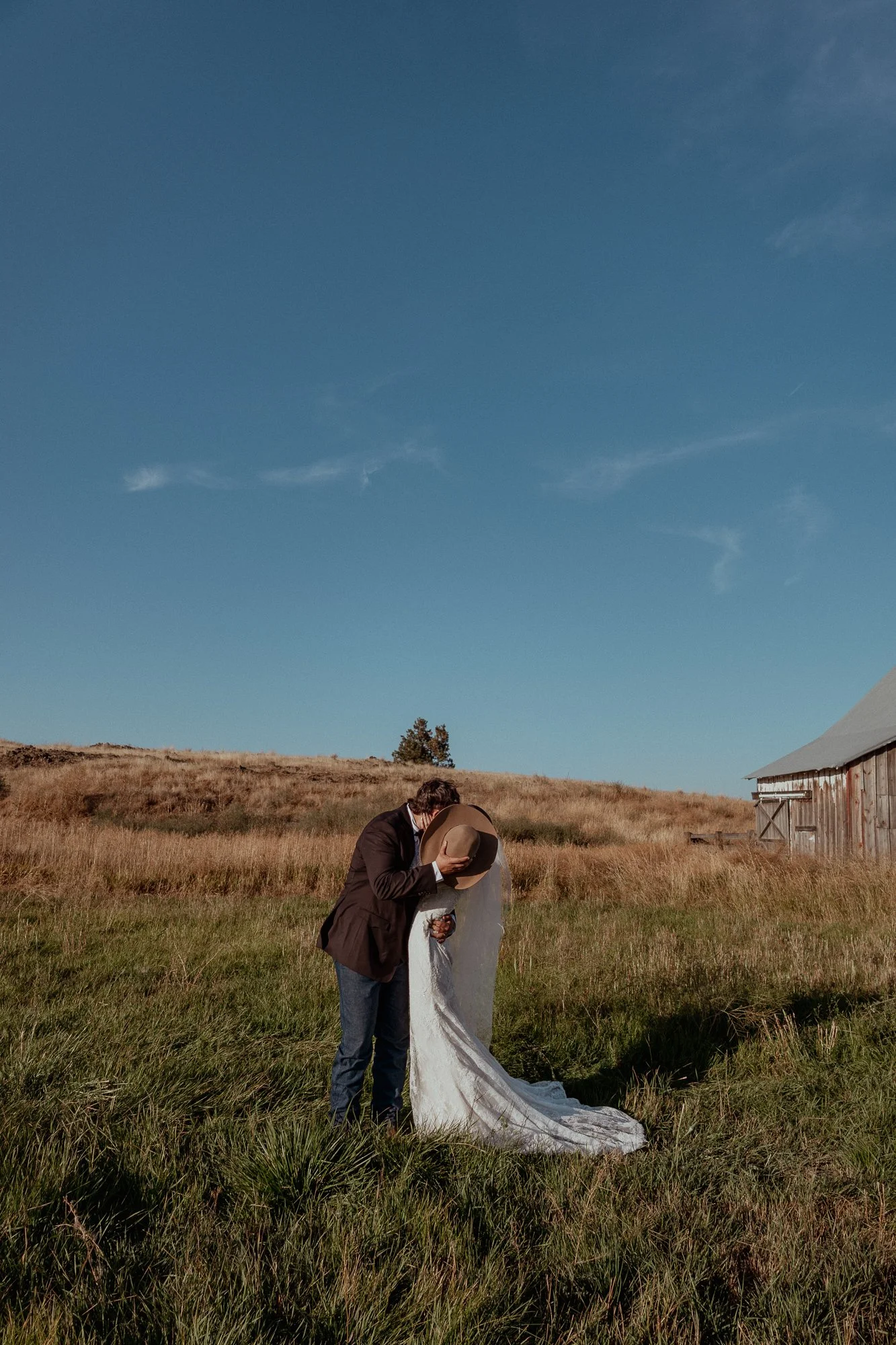 A couple dressed in wedding attire sharing a kiss in a grassy field under a clear blue sky, with a rustic barn visible on the right.