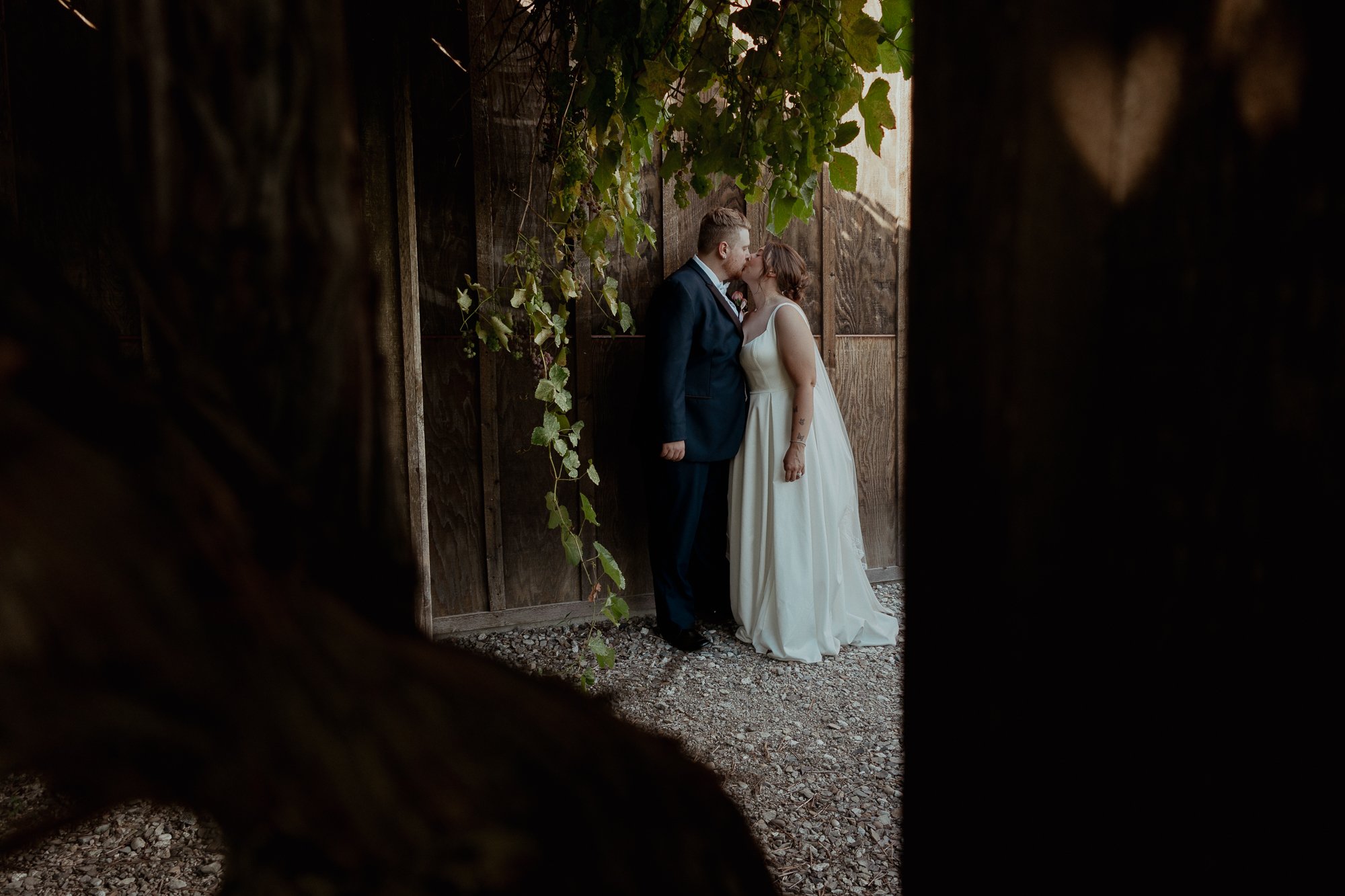 A newlywed couple shares a kiss in a rustic outdoor setting, framed by dark wooden structures and greenery.