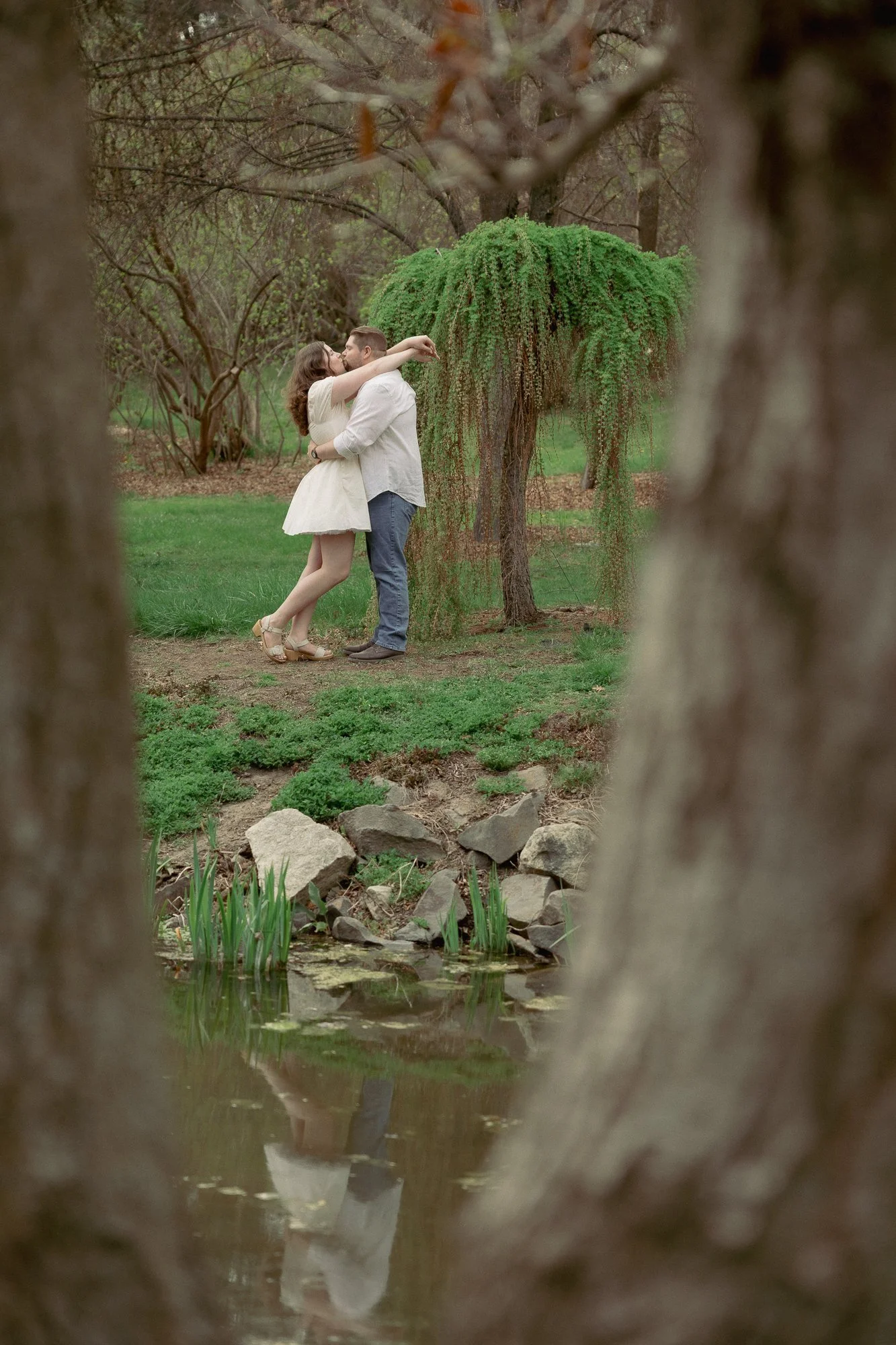 View of couple kissing next to a pond from between tree trunks
