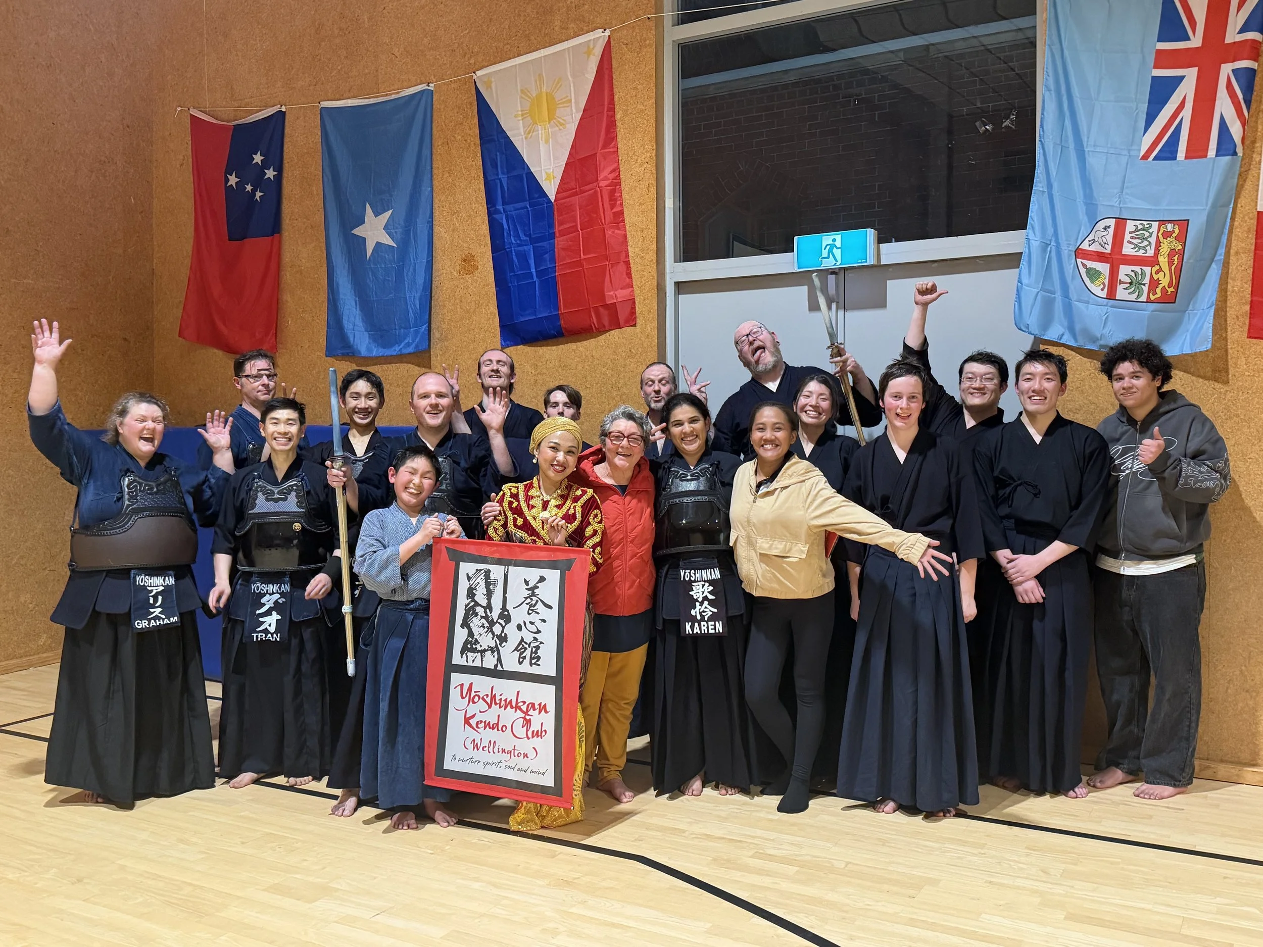 Group martial arts practice in gymnasium, featuring instructor and participants with wooden swords, wearing traditional uniforms, standing in circle.