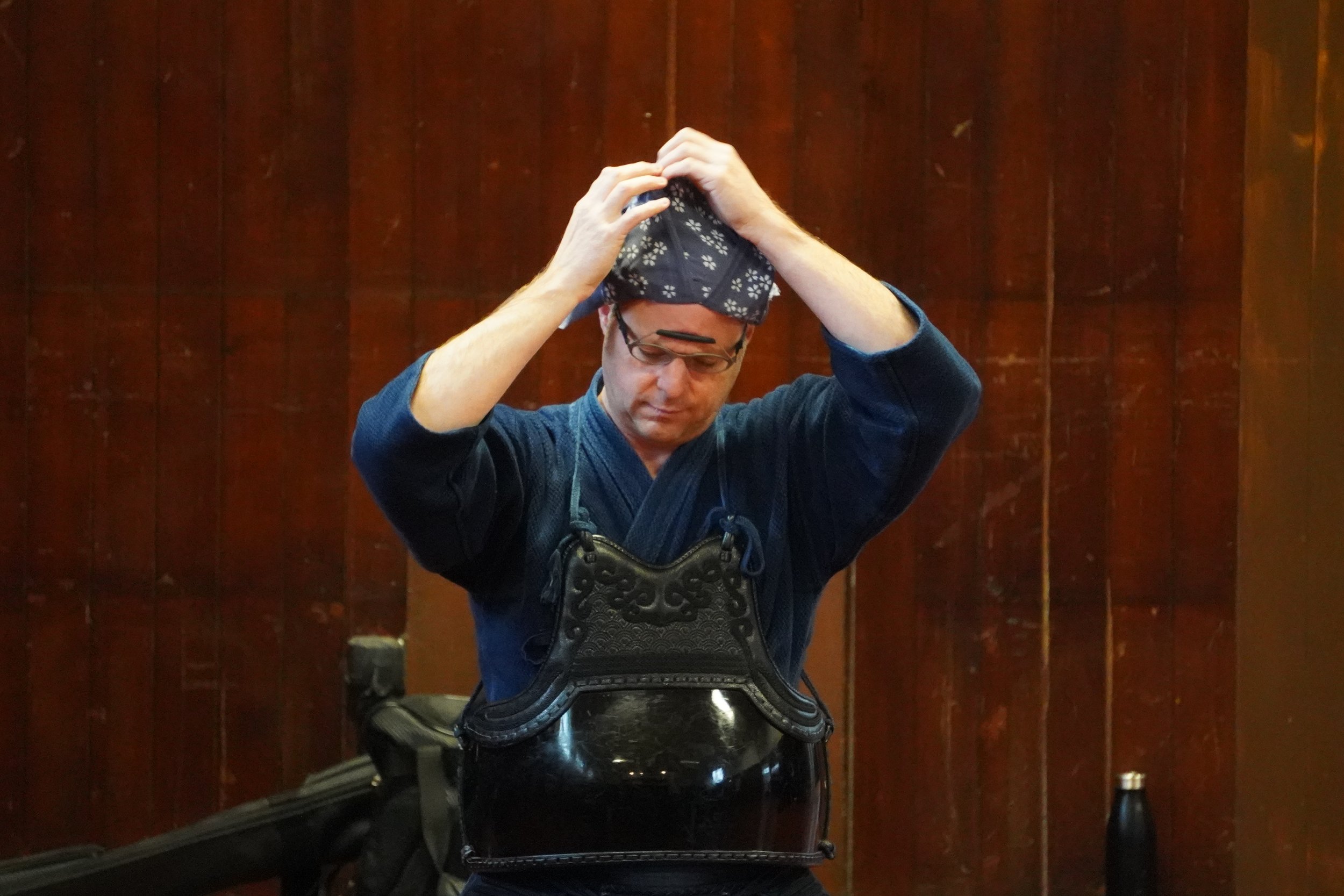 Two people practicing kendo in a gym, wearing traditional kendo armor and helmets, with one in motion holding a bamboo sword.