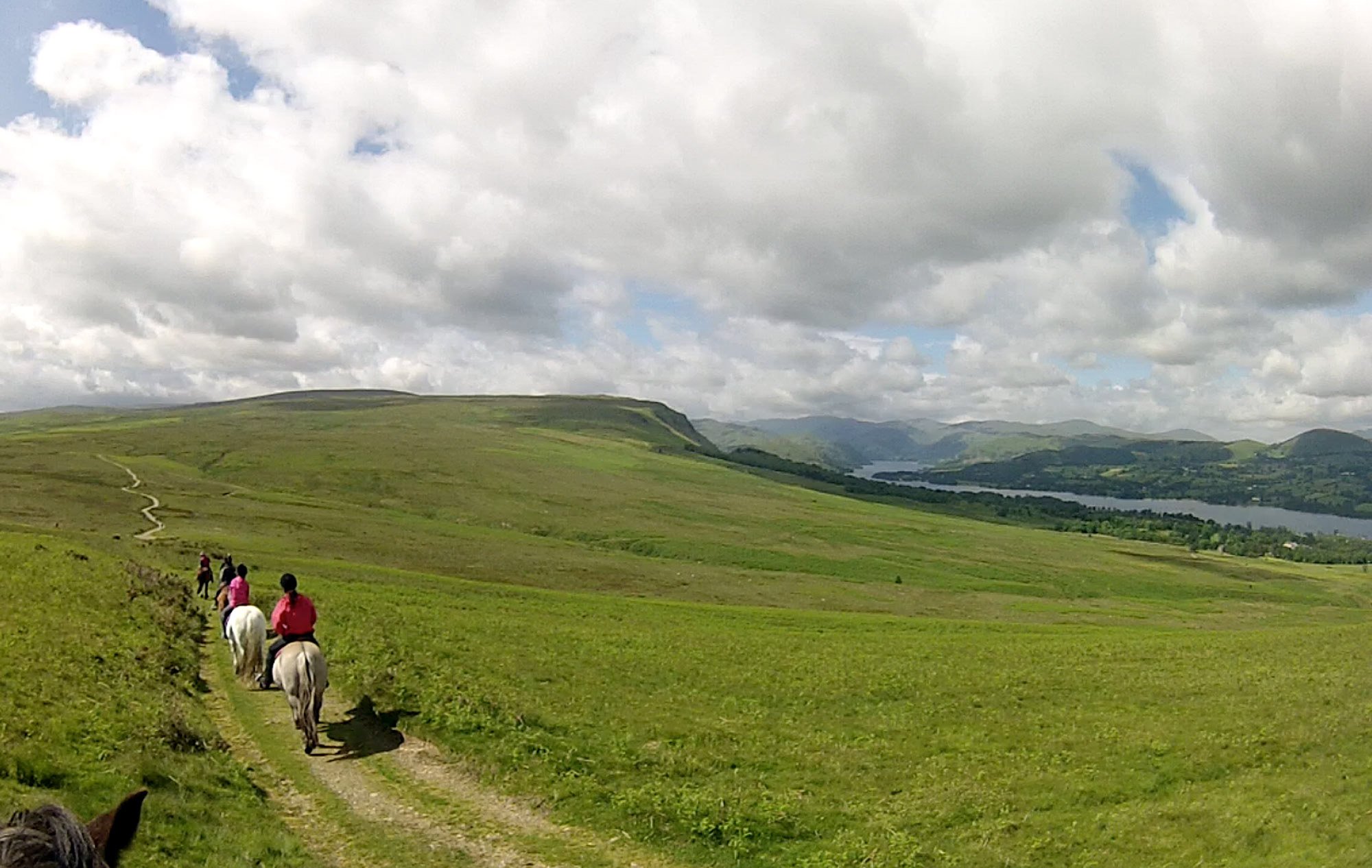 Riders on horseback traveling down a grassy trail with rolling hills, a lake, and mountains in the distance under a partly cloudy sky.