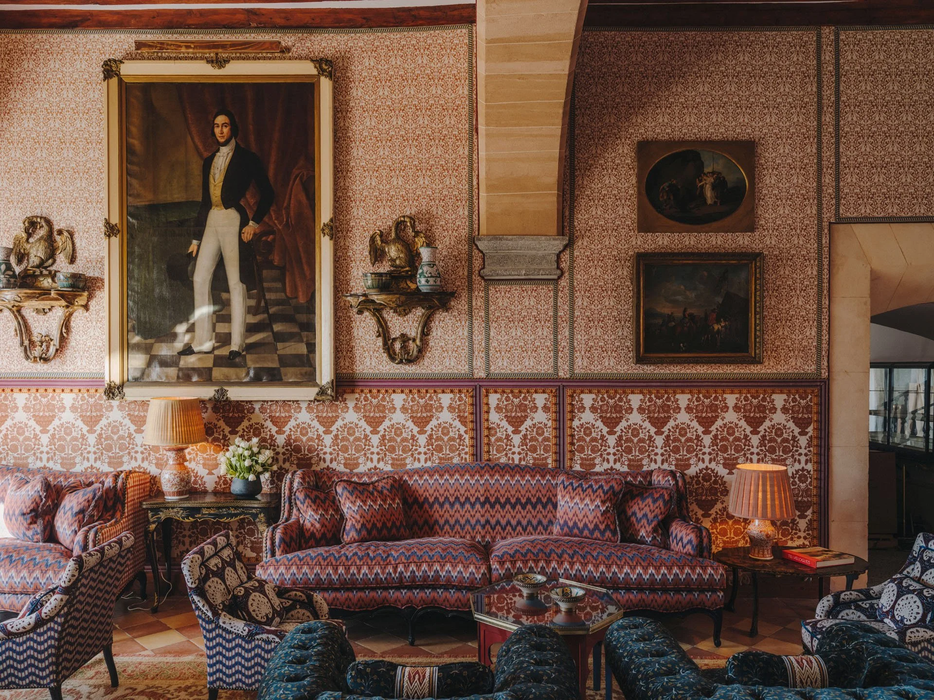 Elegant room with patterned wallpaper, a large portrait of a man, ornate wall shelves with decorative items, a sofa with multicolored zigzag pattern, chairs with geometric and floral patterns, lamps, and a small table with books and bowls.