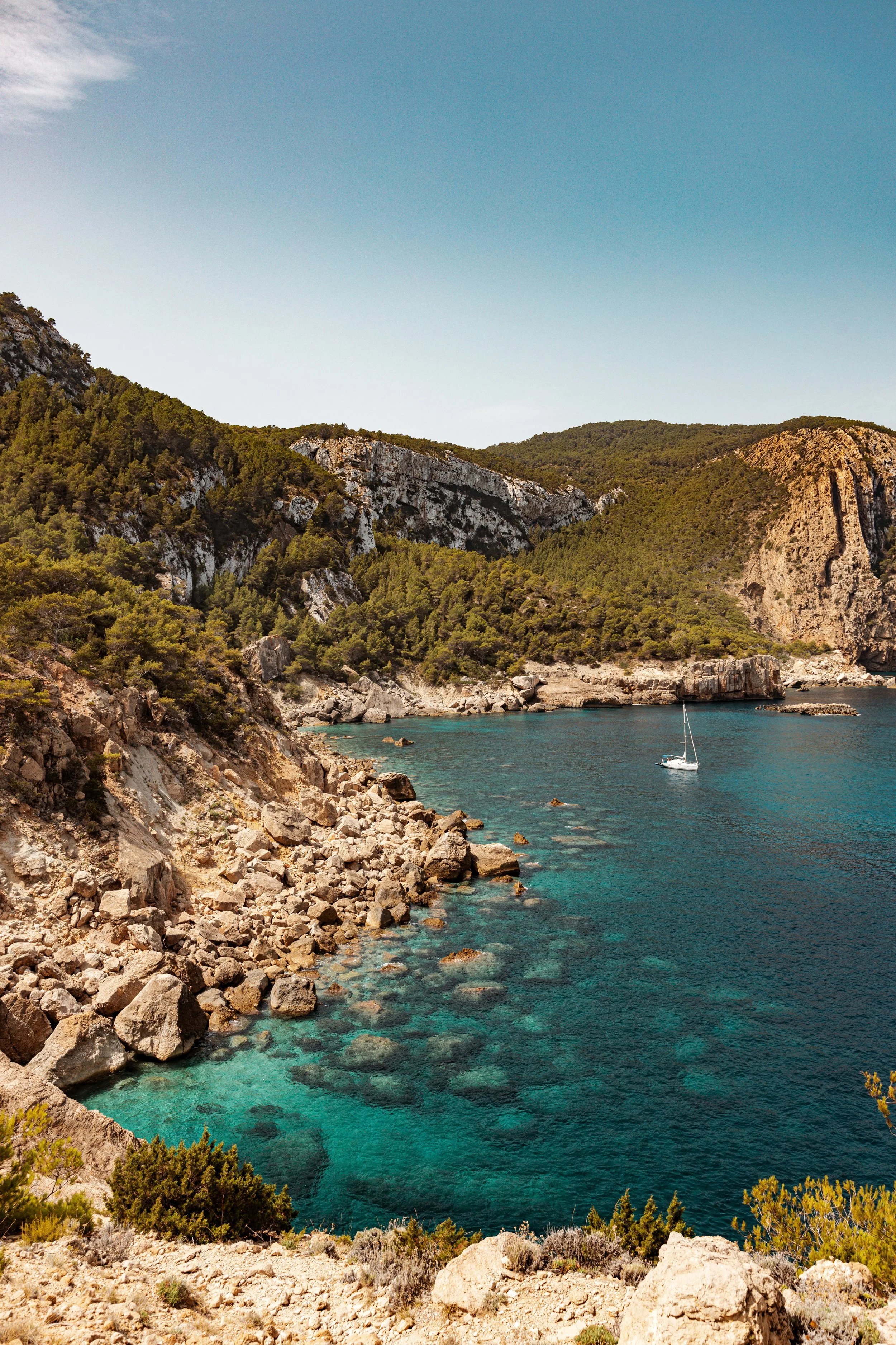 Rocky ibiza coastline with turquoise water, forested cliffs, and a sailboat in a bay.
