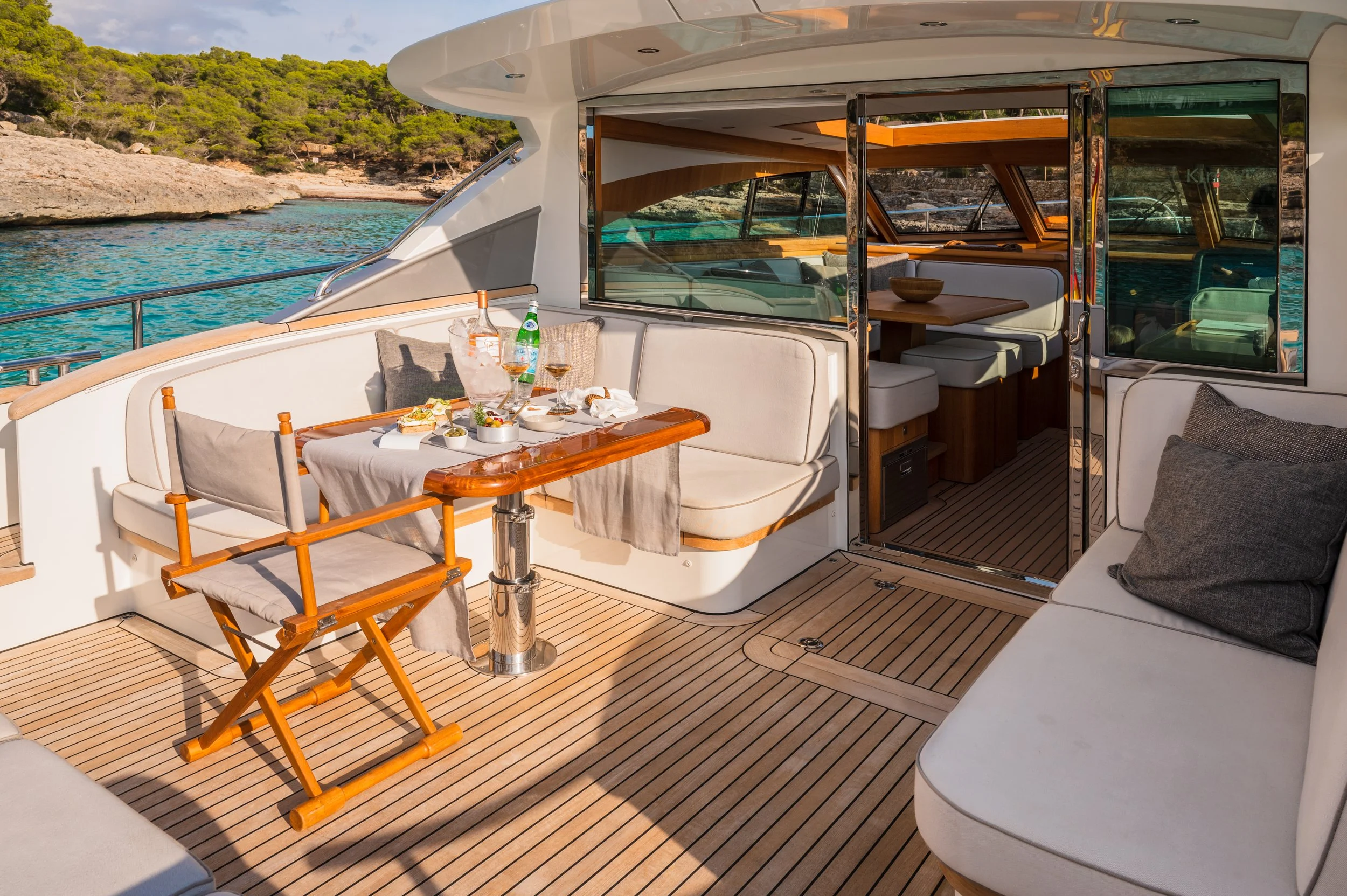 Luxury yacht deck with dining area, wooden table set with food and drinks, cushioned seating, ocean view, and interior cabin visible through glass door.