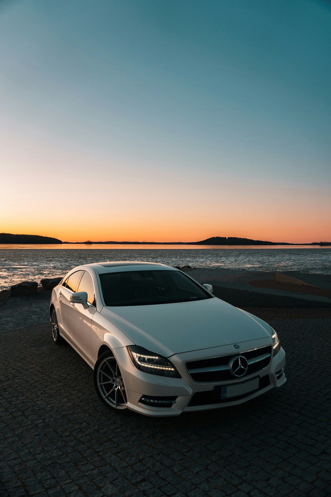 A white Mercedes-Benz sedan parked on a cobblestone area near a body of water at sunset