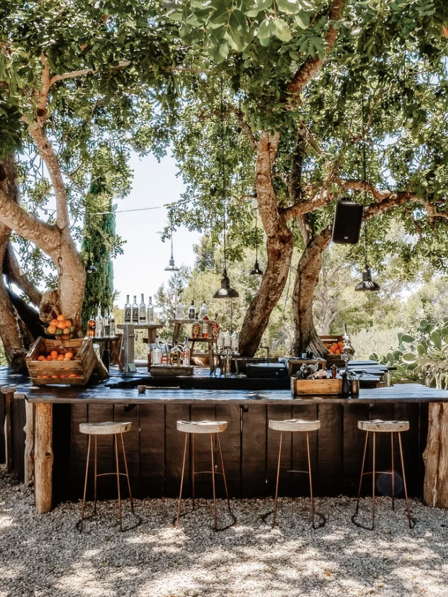 Outdoor bar under trees with wooden stools and hanging lights; bottles displayed on the counter.