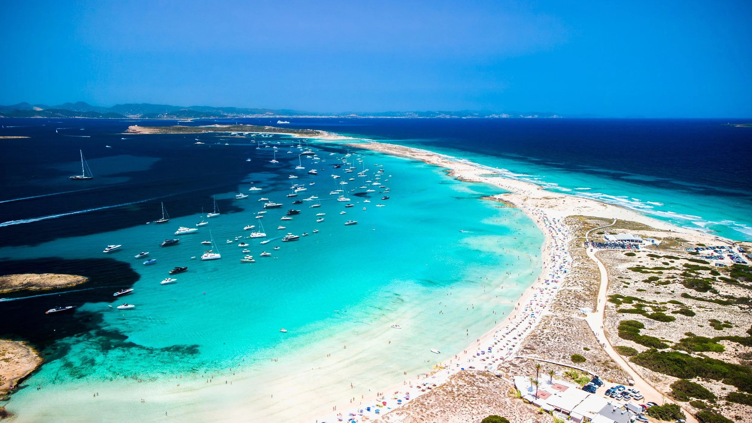 Aerial view of a turquoise beach in formentera with boats anchored and a sandy shoreline, featuring people and umbrellas.