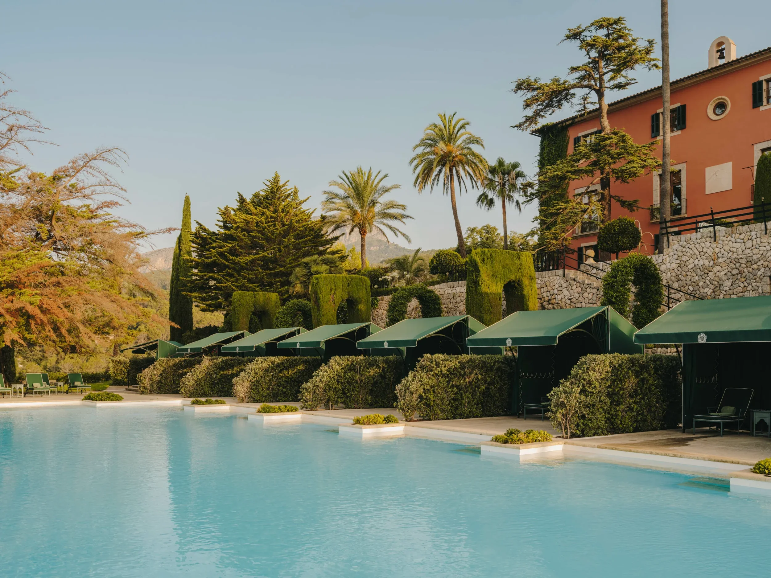A luxurious outdoor swimming pool surrounded by green cabanas and lush landscaping with tall palm trees and a pink building in the background.