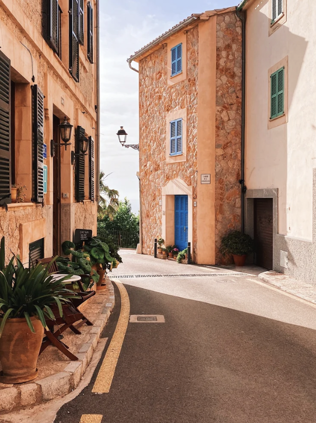 Narrow mallorcan street with stone buildings, blue shutters, and potted plants, under a clear sky.
