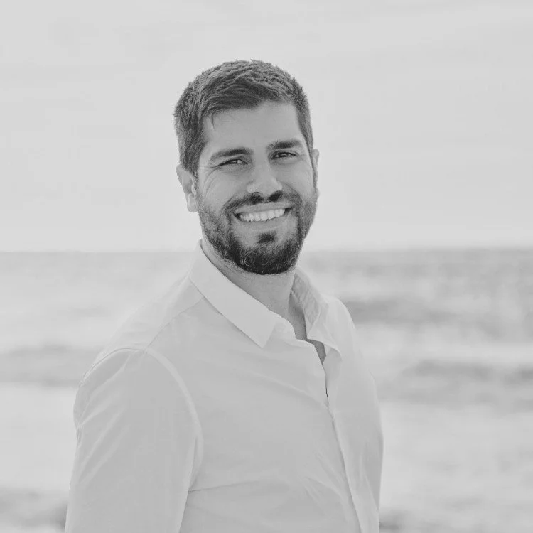 A smiling man with short hair and a beard, wearing a light-colored button-up shirt, standing on a beach with the ocean in the background.