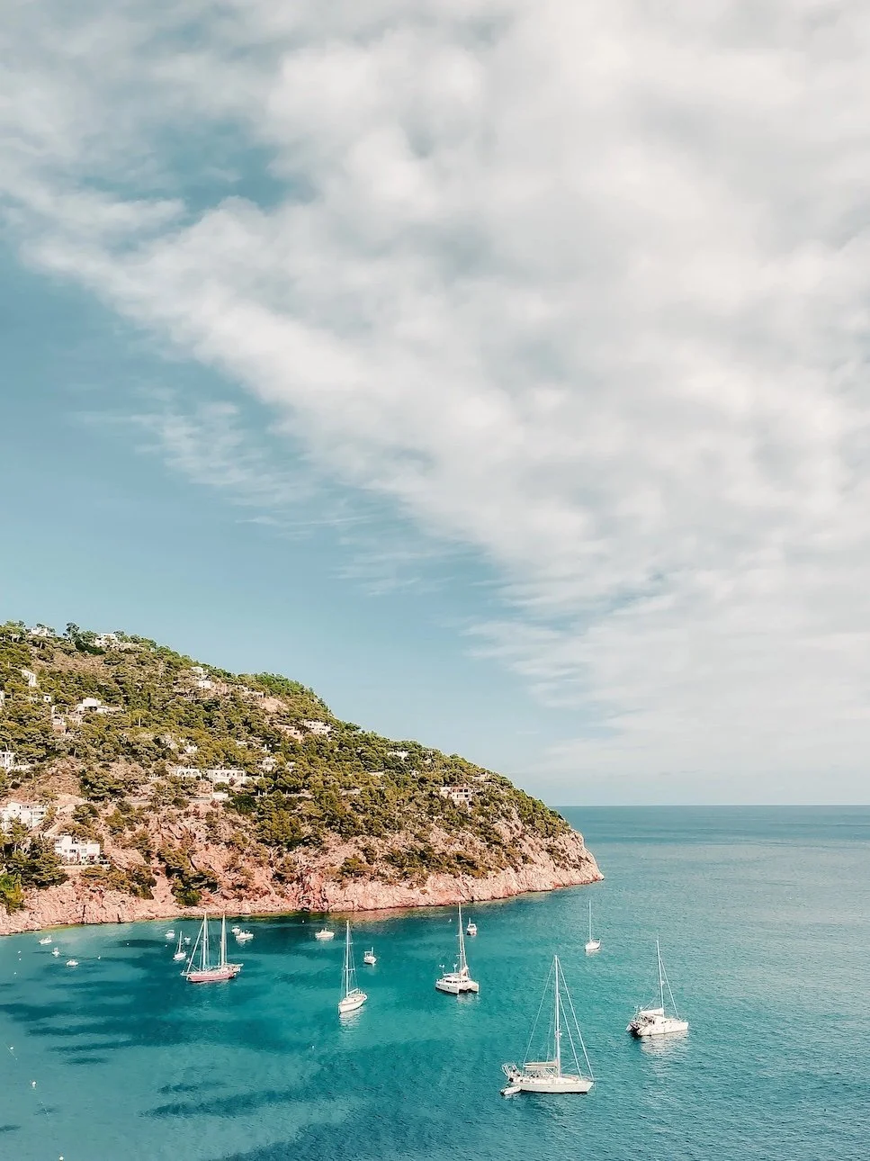 View of a coastal hillside with houses and trees overlooking a turquoise bay with sailboats