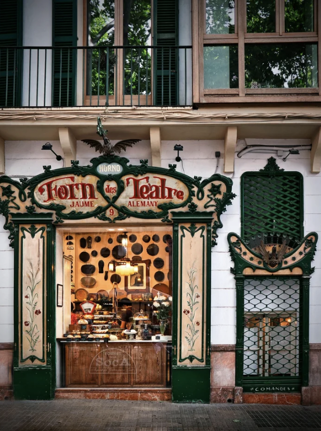 An ornate bakery entrance with decorative green and beige elements, displaying the sign "Forn des Teatre Jaume Alemany." A display window shows a variety of baked goods inside. The building facade features windows with shutters and wooden frames above the entrance.