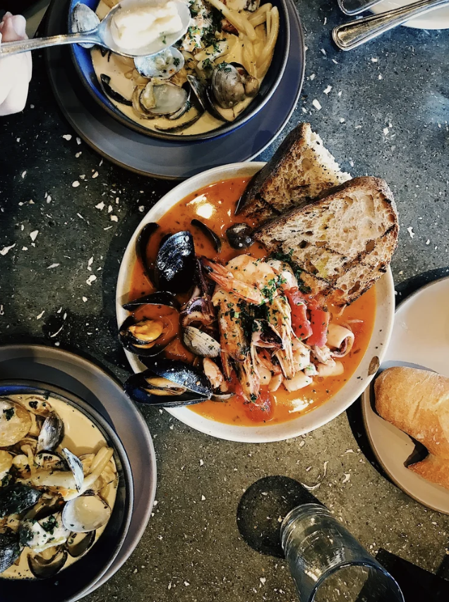 Seafood pasta dishes with clams, mussels, shrimp, toasted bread; glass of water on a table.
