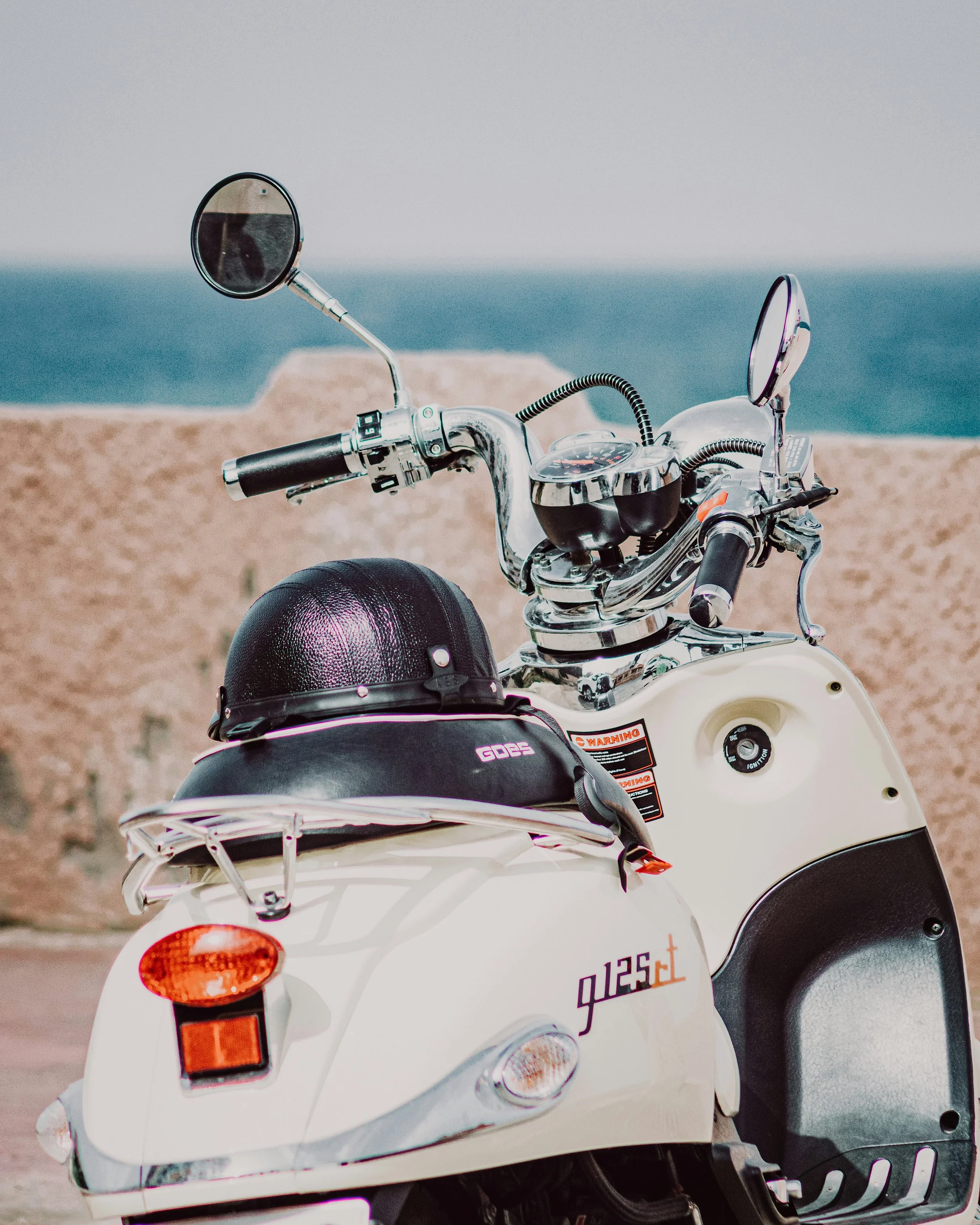 A white scooter parked near the coast with a large rocky island and ocean in the background.
