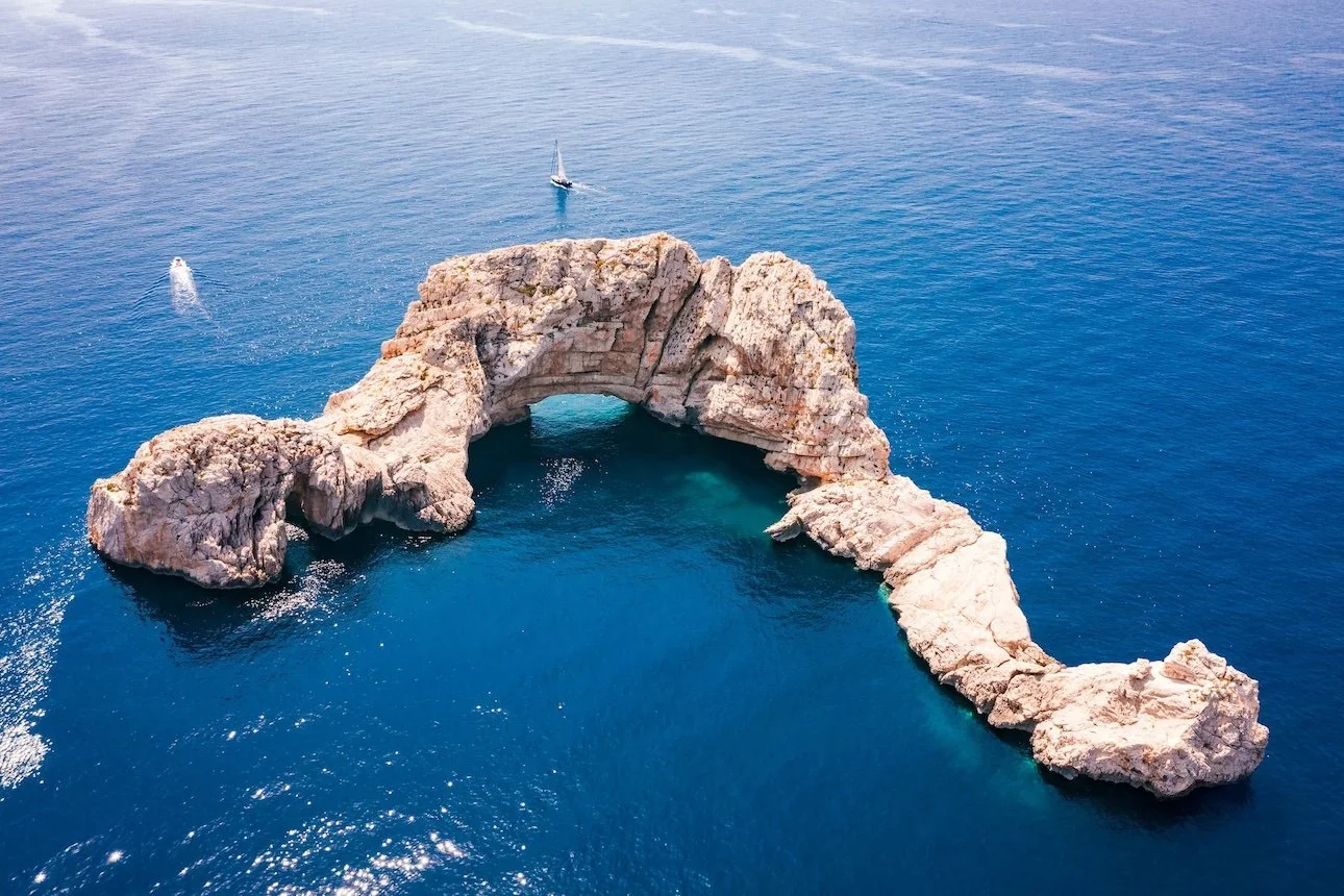 A large rock formation in the shape of an arch with a hole in the middle, situated in the ocean, with a sailboat nearby.