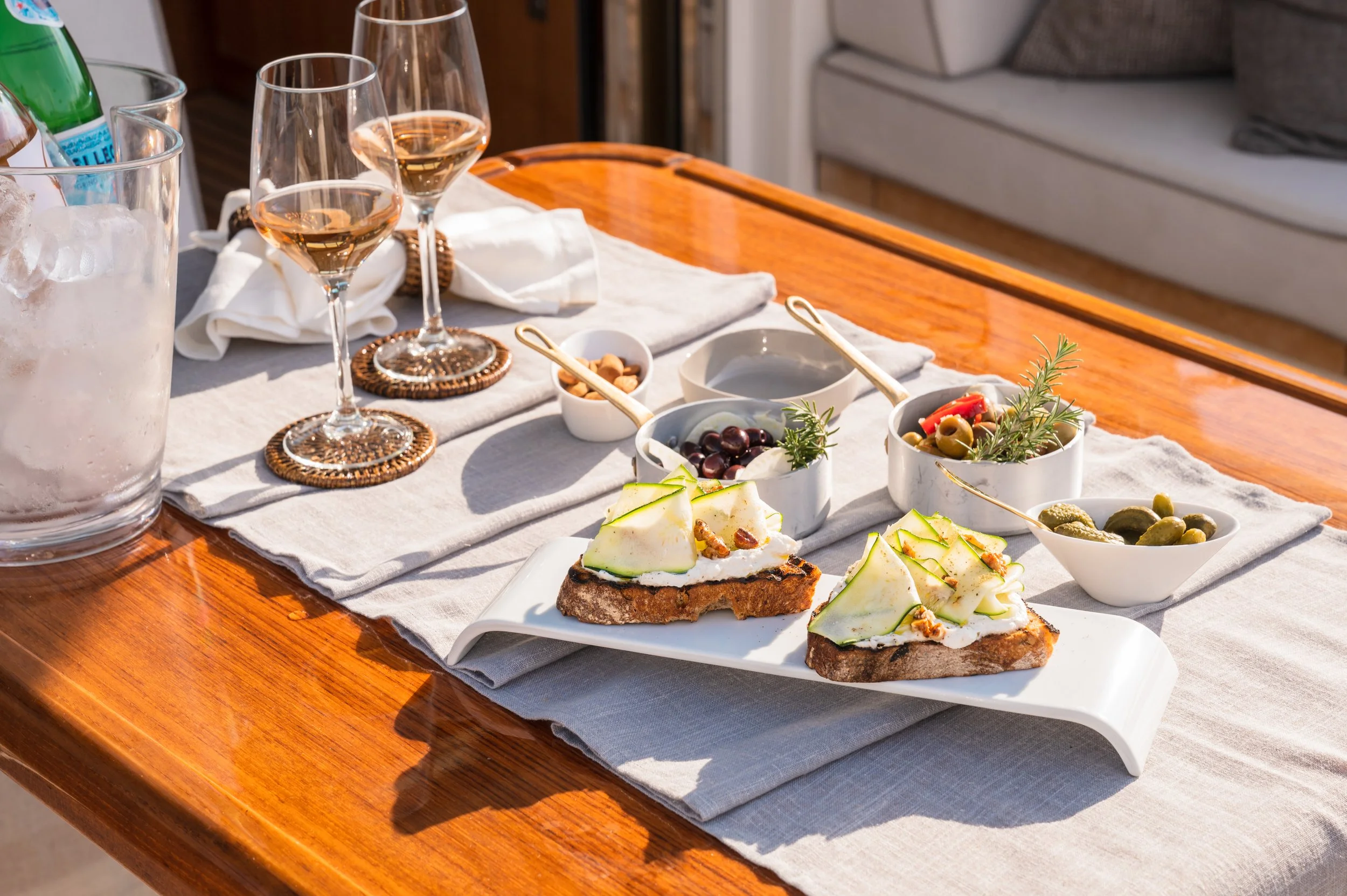 Outdoor dining setup with wine glasses, bruschetta, and small bowls of olives, nuts, and pickles on a wooden table with a linen runner