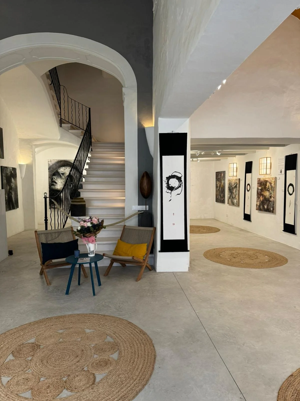 Interior of an art gallery with paintings on the walls, a staircase with a black wrought iron railing, two wooden chairs with pillows and a small table with a bouquet of flowers.
