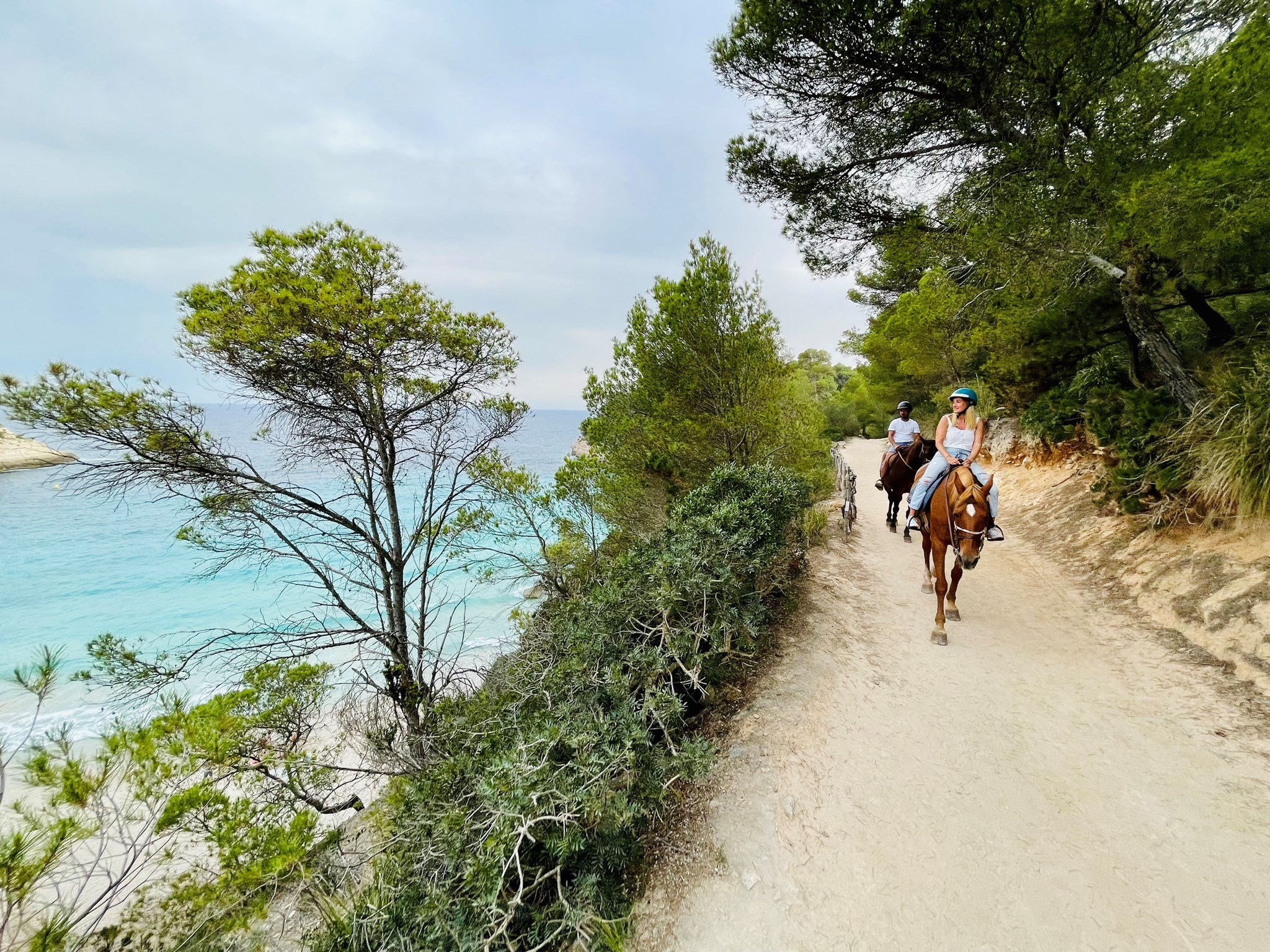 Three people riding horses along a dirt trail near the coast, with trees and bushes on one side and the ocean on the other.