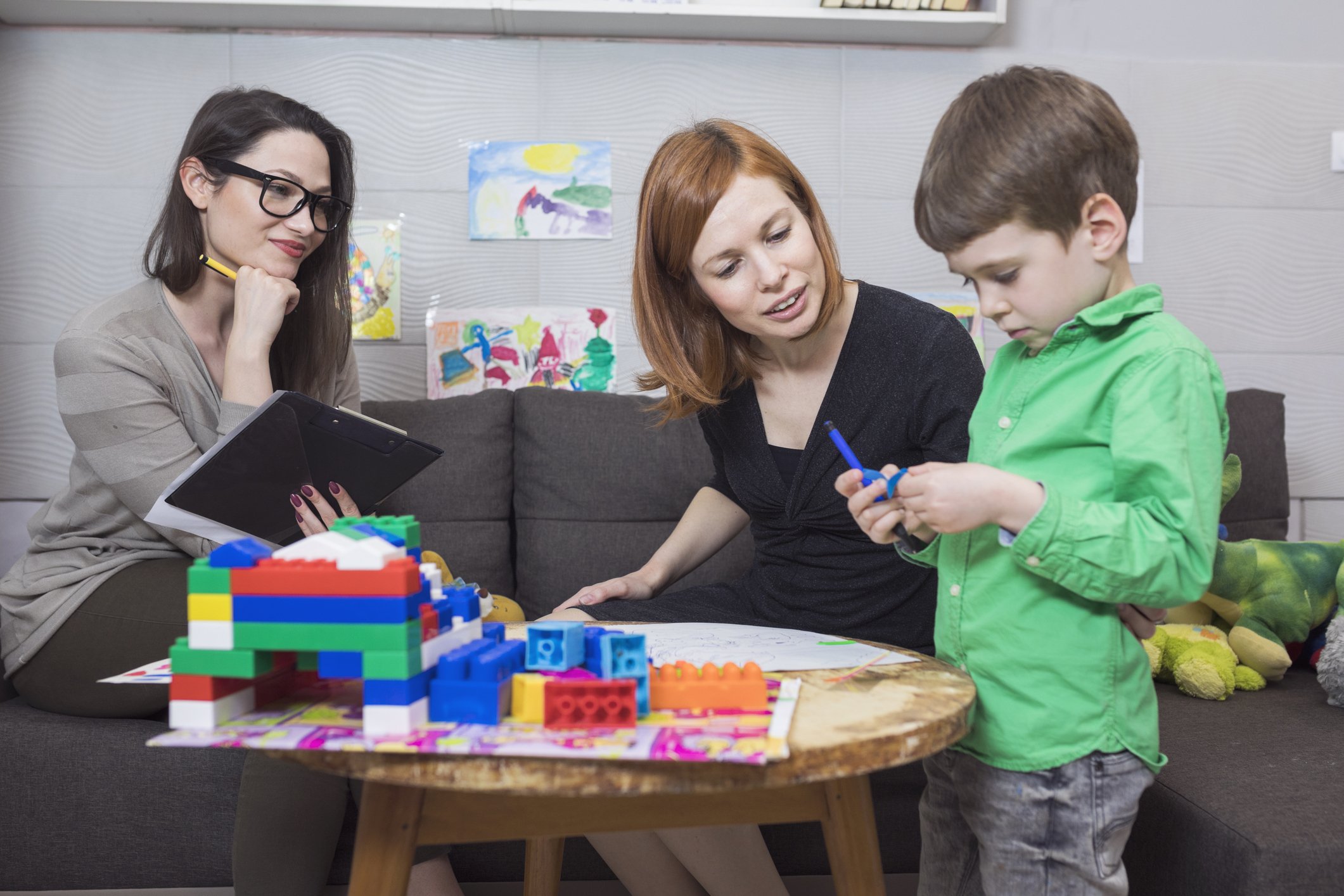 Child psychologist counseling young boy in therapy session with toys and drawings on display.