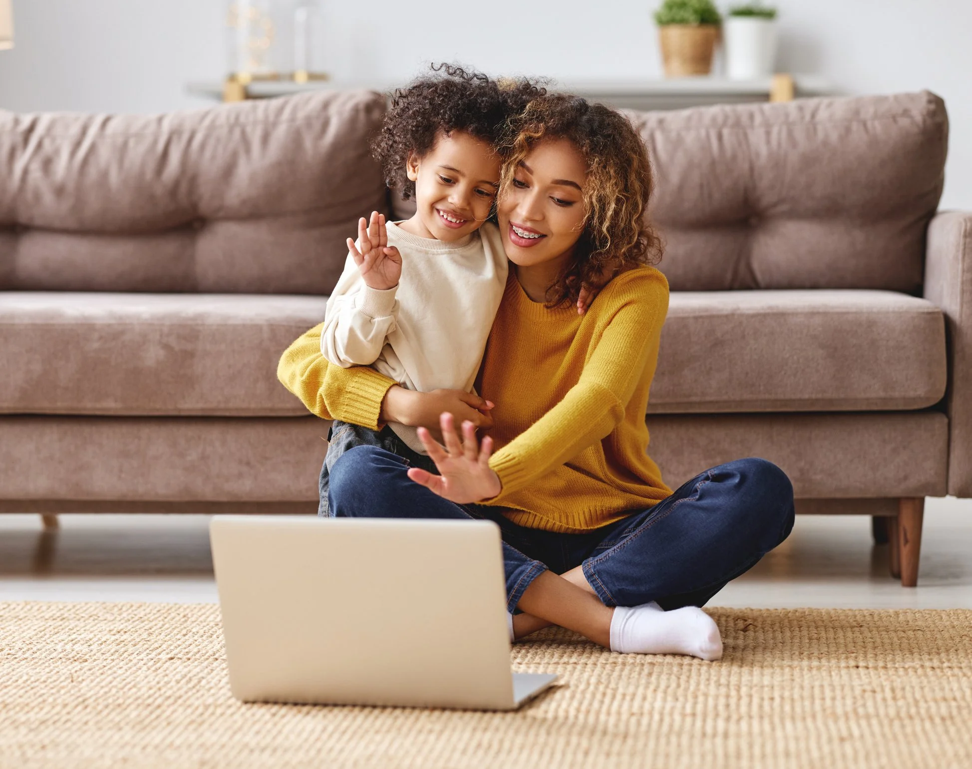 A person and a child sitting on the floor in front of a laptop, waving at the screen. They are in a cozy living room with a sofa in the background.