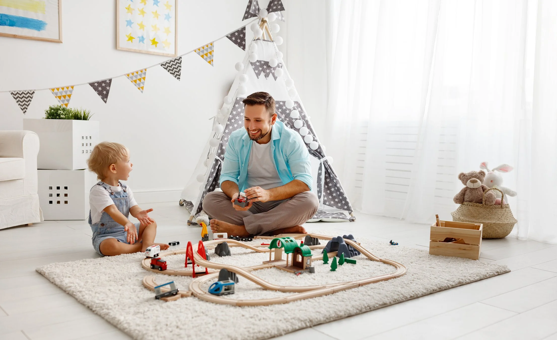 A child and an adult playing with a toy train set on a carpet in a bright room, with a teepee, framed art, and stuffed animals in the background.