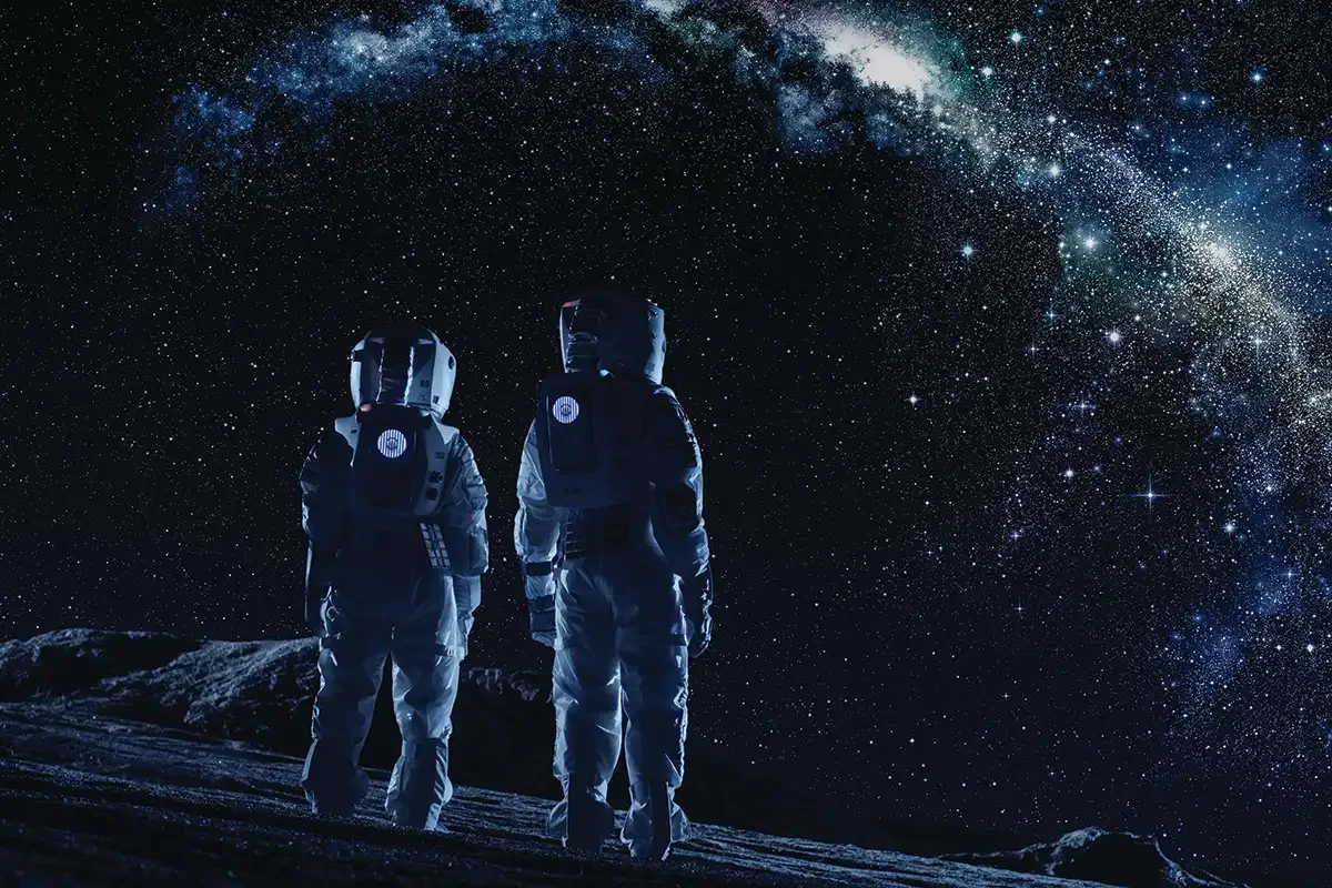 Two astronauts in spacesuits standing on the moon's surface, gazing at a star-filled sky and the Milky Way galaxy.