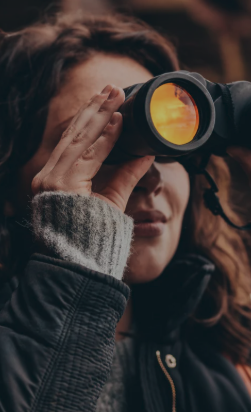 Person looking through binoculars outdoors, smiling, wearing a gray sweater and black jacket.