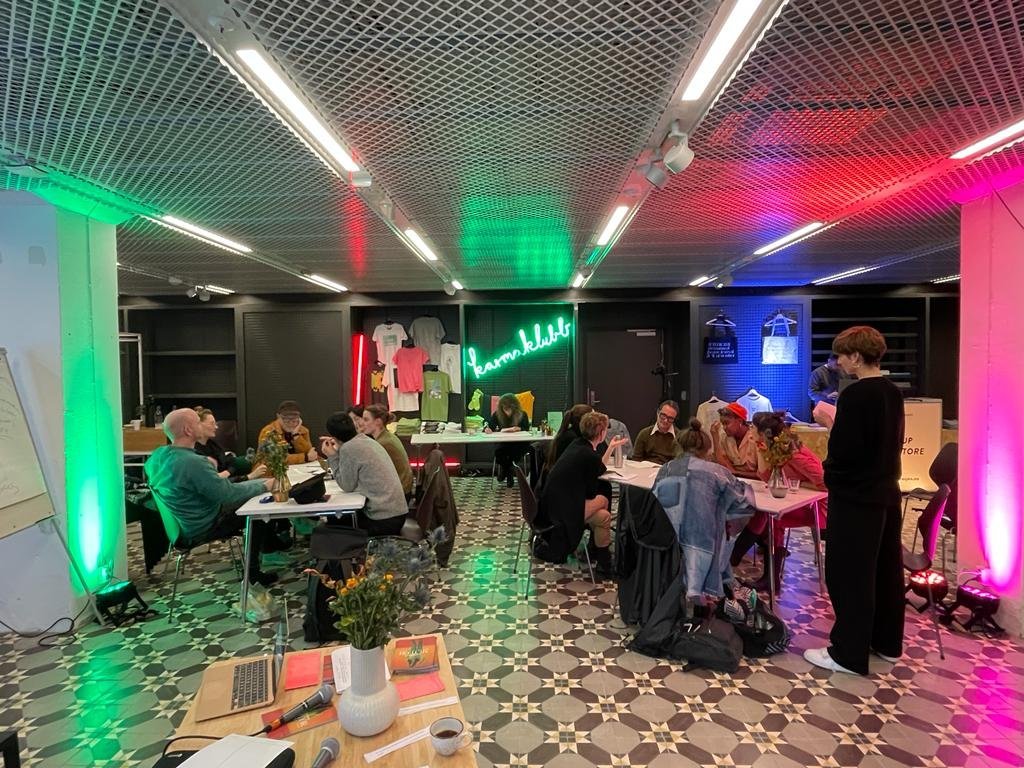  Open room. Groups working at tables. Black, white patterned floor, neon lights. 