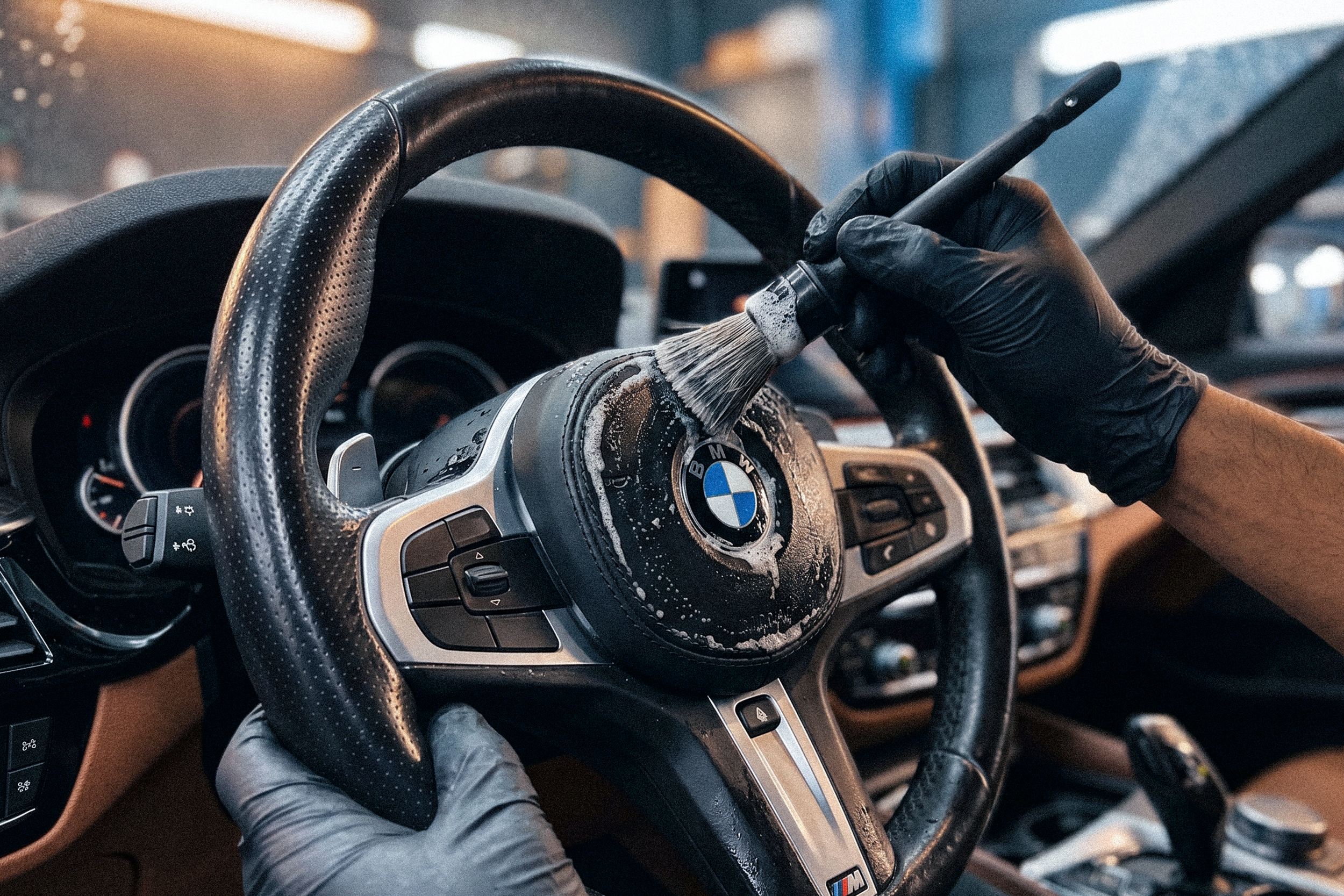 A person wearing black gloves cleaning the steering wheel of a BMW car with a brush and soap foam.