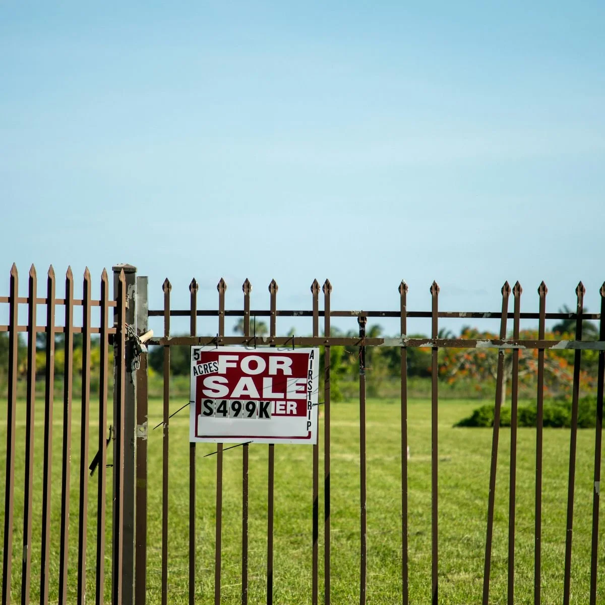 blue sky green grass and an iron fence around land with a for sale sign
