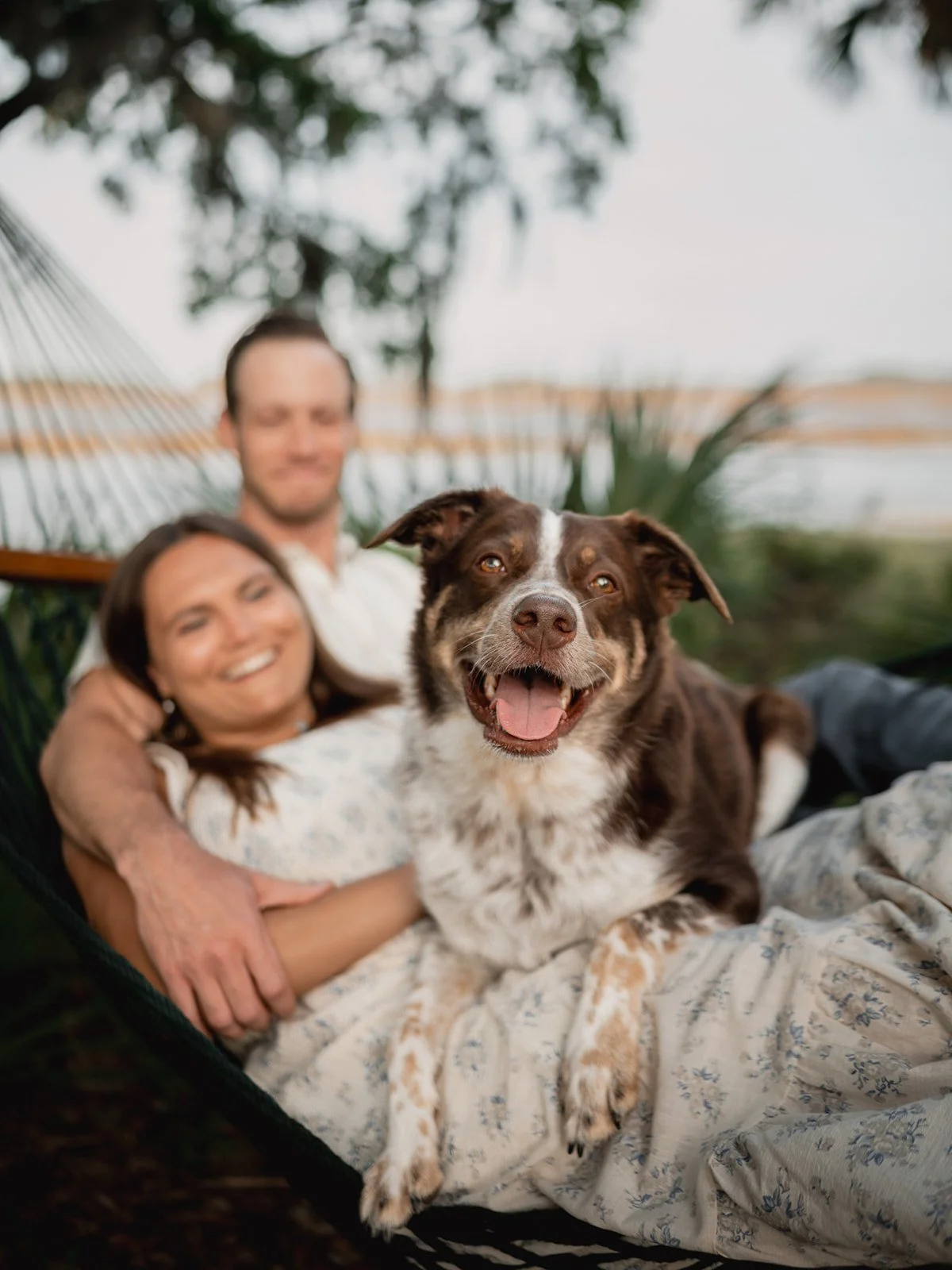 Couple lounging in a hammock with their dog between them, smiling and relaxed.
