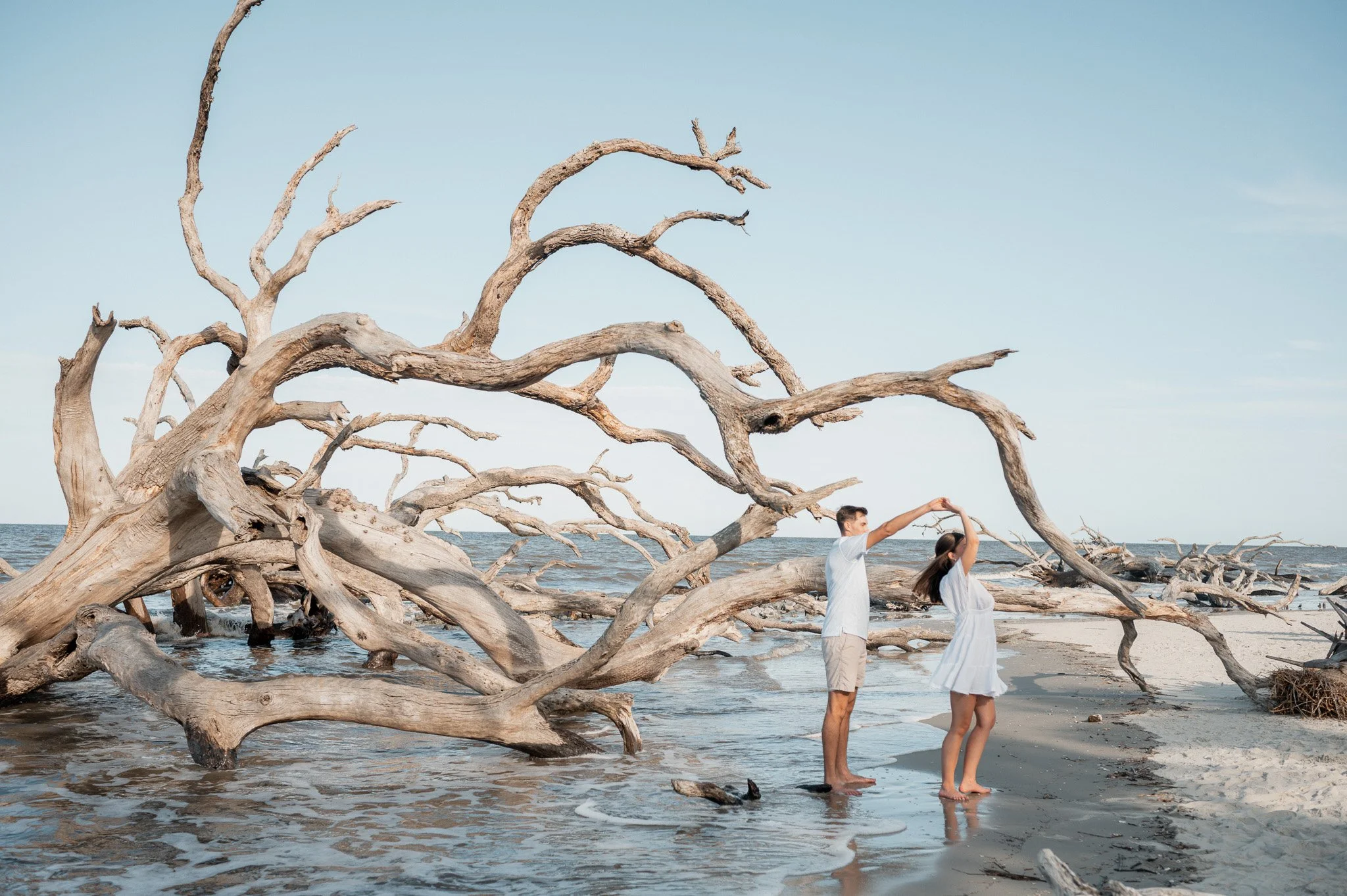 Couple holding hands during Hunting Island engagement shoot on driftwood beach at sunset, with ocean waves and weathered tree trunks behind them.