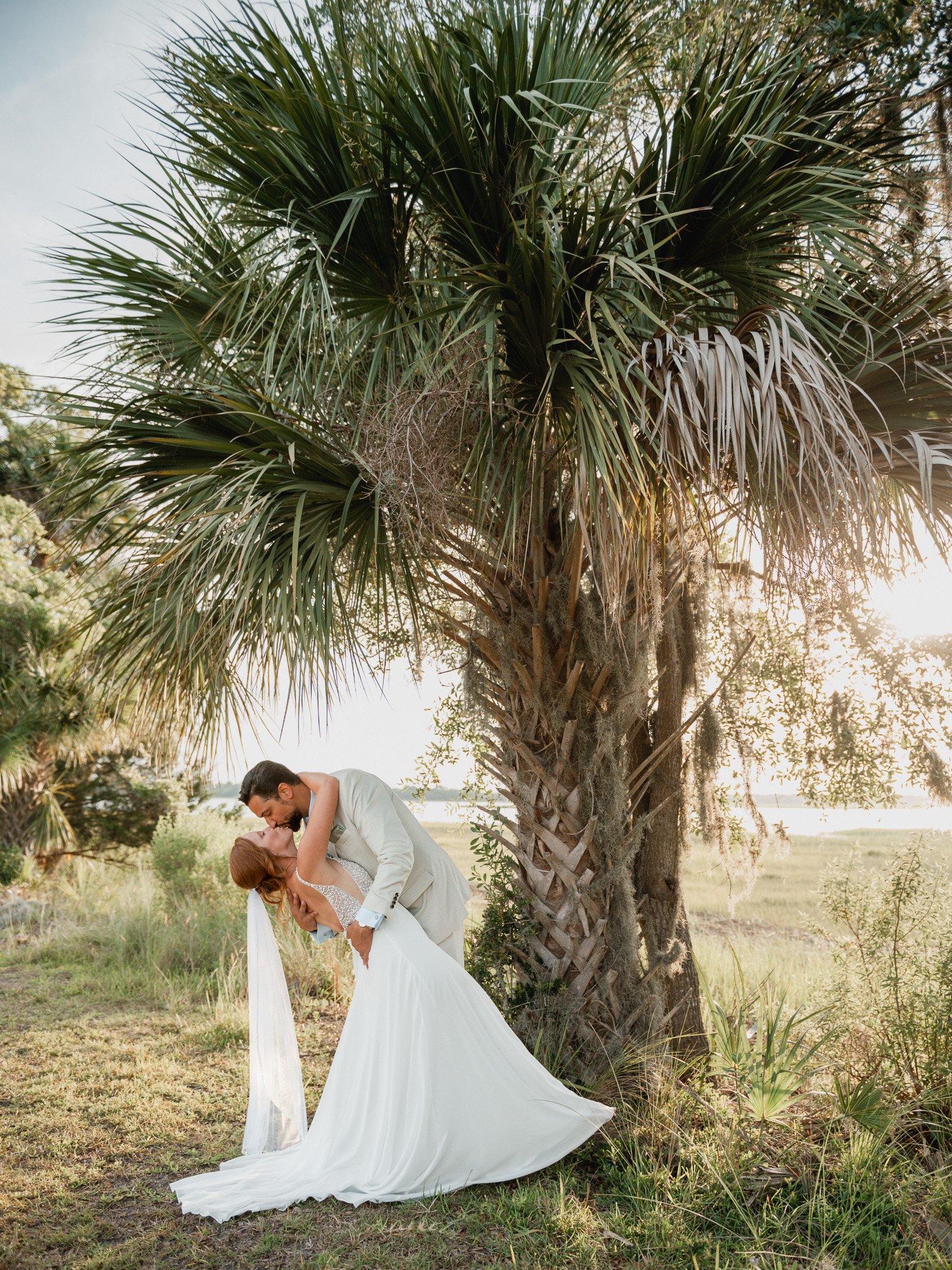 Bride and groom standing under a palm tree, husband dipping and kissing his bride after a micro‑wedding ceremony on a sunny day.