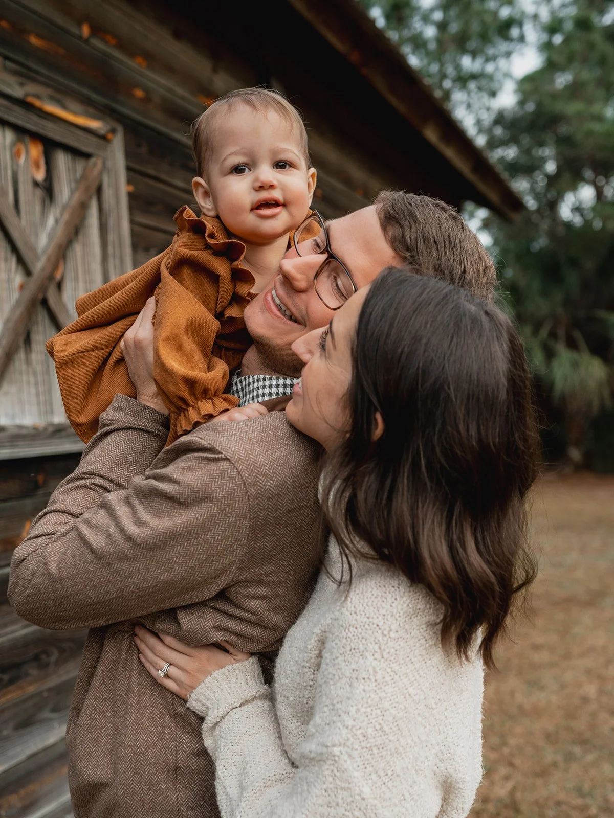 Parents hugging while their toddler wraps arms around them from behind, all smiling brightly.