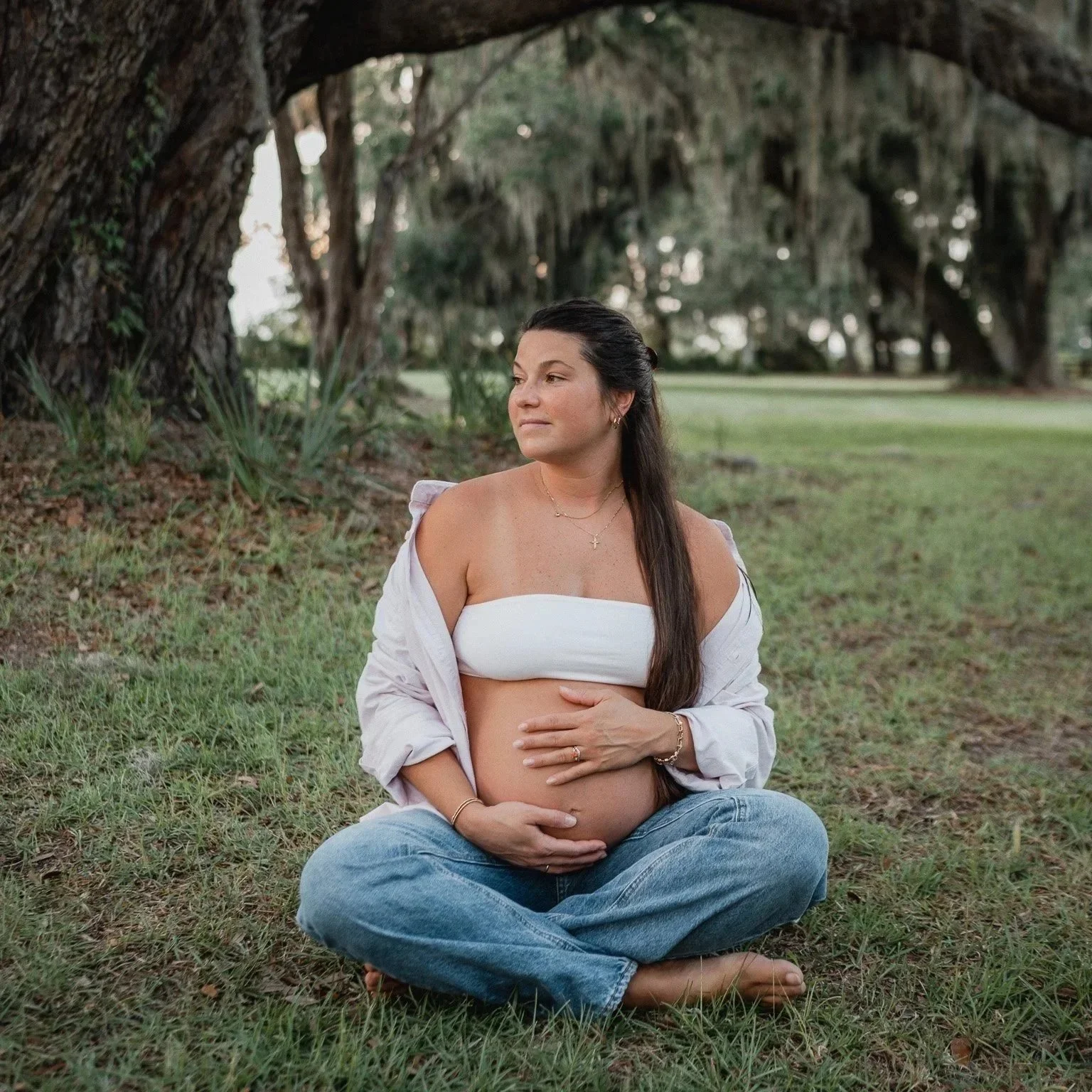 Expectant mother sitting cross‑legged under a sprawling Lowcountry oak tree, gently holding her baby bump. Maternity photo took place in riverside park in Beaufort, SC.